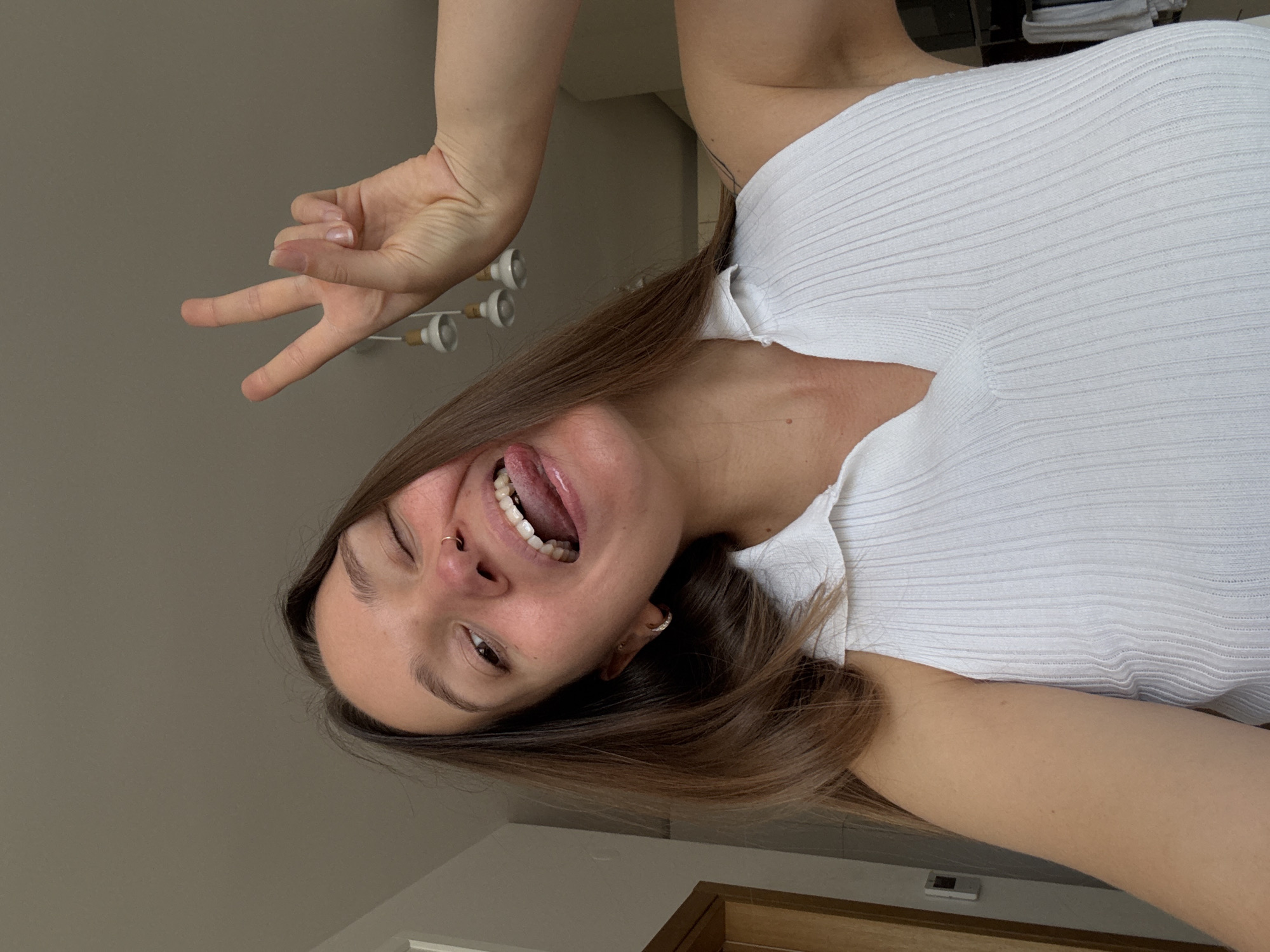 A young woman with long brown hair is taking a selfie. She is wearing a white ribbed halter top and has her tongue sticking out, winking with her right eye. Her left eye is open. She is making a peace sign with her right hand. The background is an indoor setting with neutral-colored walls and ceiling. There are three pendant lights visible in the background. A door frame is visible on the left side of the image. The woman has a nose ring and hoop earrings. The overall impression is a casual and playful moment captured in an indoor environment. The location of Boreti, Montenegro is not indicated by any visual cues in the image.