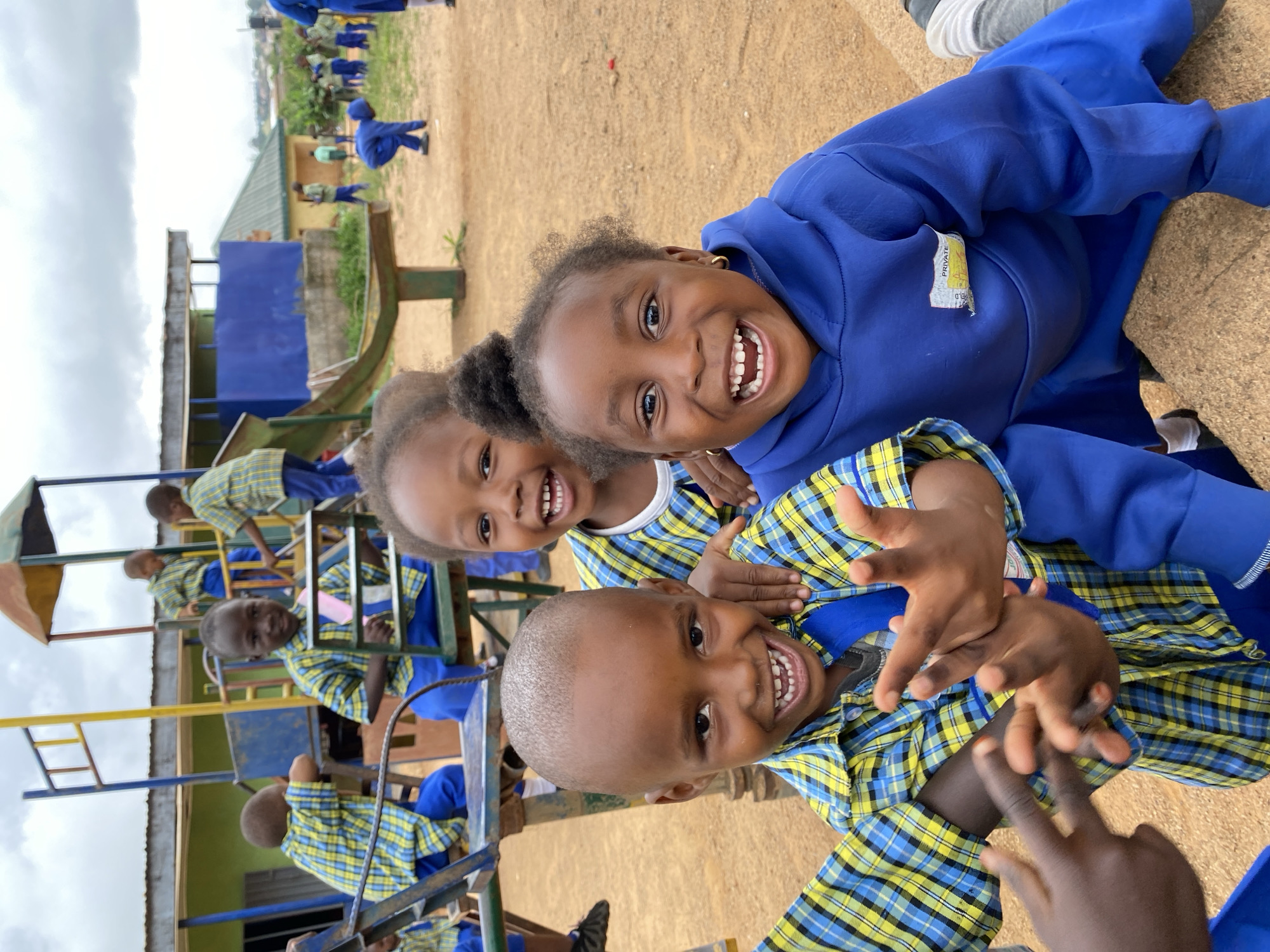 Three children are pictured outdoors in Gura Top, Nigeria. They are all smiling and appear to be playing together. Two of the children are wearing a blue and yellow plaid uniform shirt with a blue undershirt. The third child is wearing a blue hooded sweatshirt. They are positioned close together, possibly embracing or posing for a photo. In the background, there's playground equipment, and other children in blue uniforms can be seen, suggesting they are at a school or recreational area. The ground is dirt or sand. The overall environment suggests a sunny day at a playground.