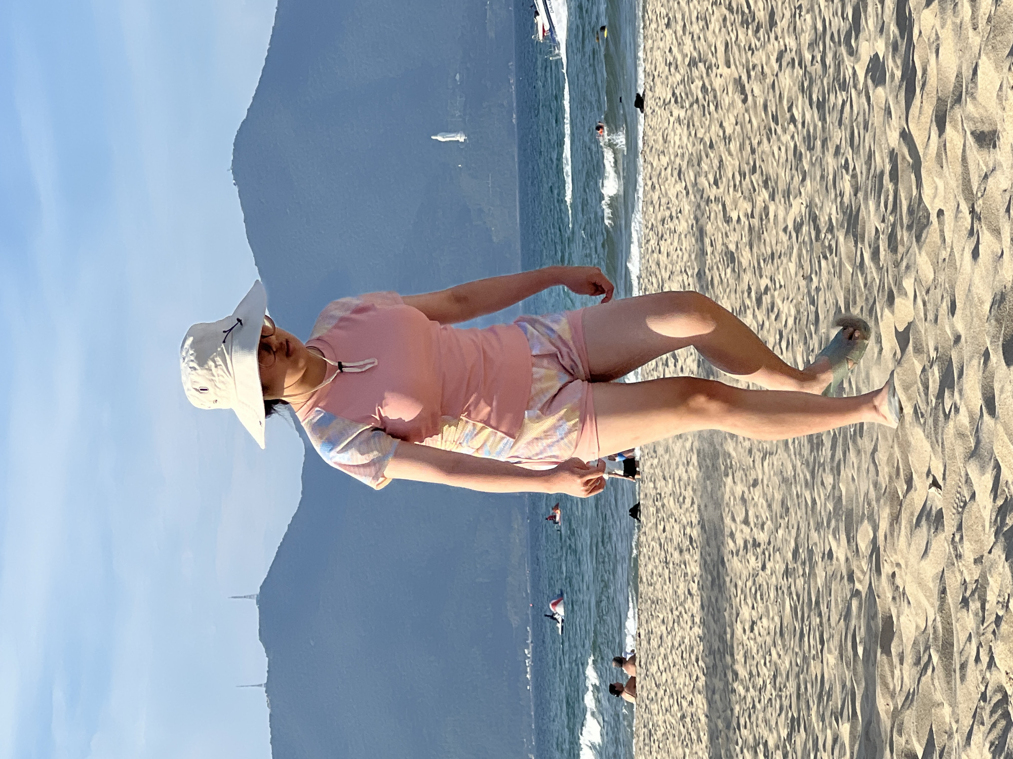 This is a vertical, full-shot photograph taken on a sunny day at a beach. The main subject is a young person, likely female, wearing a white sun hat, a pink t-shirt with a tie-dye pattern on the sleeves and collar, and patterned shorts. The person is sitting on the sand, with their legs extended towards the water. They are looking down and holding a small white object, possibly a cigarette, in their right hand.
The background shows a sandy beach with clear blue water and gentle waves. Several other people are visible in the distance, some in the water and some walking on the beach. A white boat is also visible in the water. In the far background, a mountain range is silhouetted against a clear blue sky. Two tall structures, possibly antennas or towers, are visible on the mountain.
The lighting suggests it is daytime, likely mid-morning or late afternoon, given the shadows cast on the sand. The overall mood of the image is relaxed and summery. The location is identified as Đà Nẵng, Vietnam, which is consistent with the beach and mountain scenery. There is no visible text in the image.