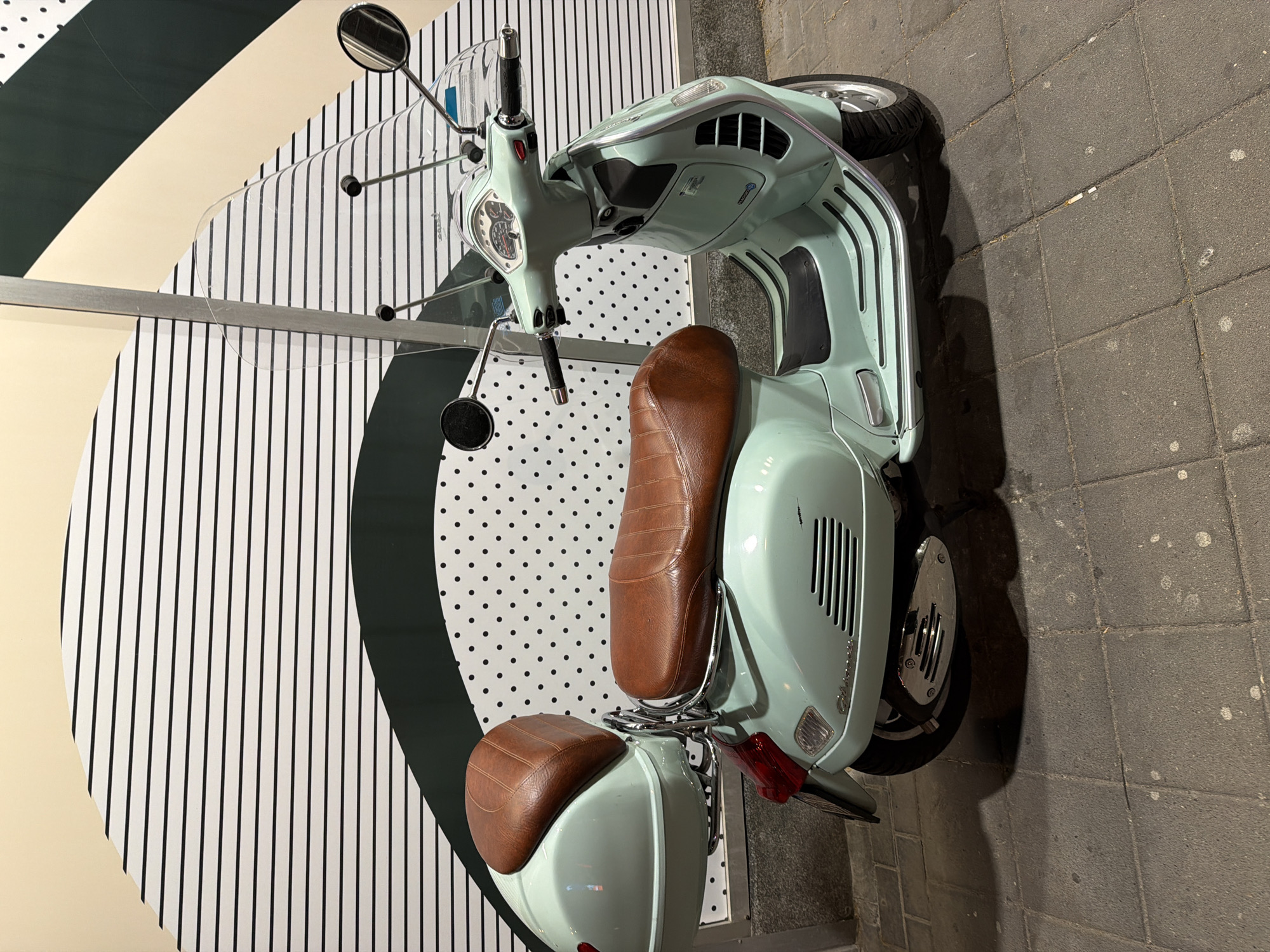 The image shows a light blue Vespa scooter parked on a gray brick pavement, seemingly outdoors. The scooter has a brown leather seat. Behind the scooter is a decorative wall with a circular pattern featuring vertical black and white stripes, a black band, and a section with white dots on a dark background. The wall appears to be part of a shop or display. The image does not contain enough information to determine the exact location in Vracar Urban Municipality, Serbia, or any specific people, actions, or interactions.