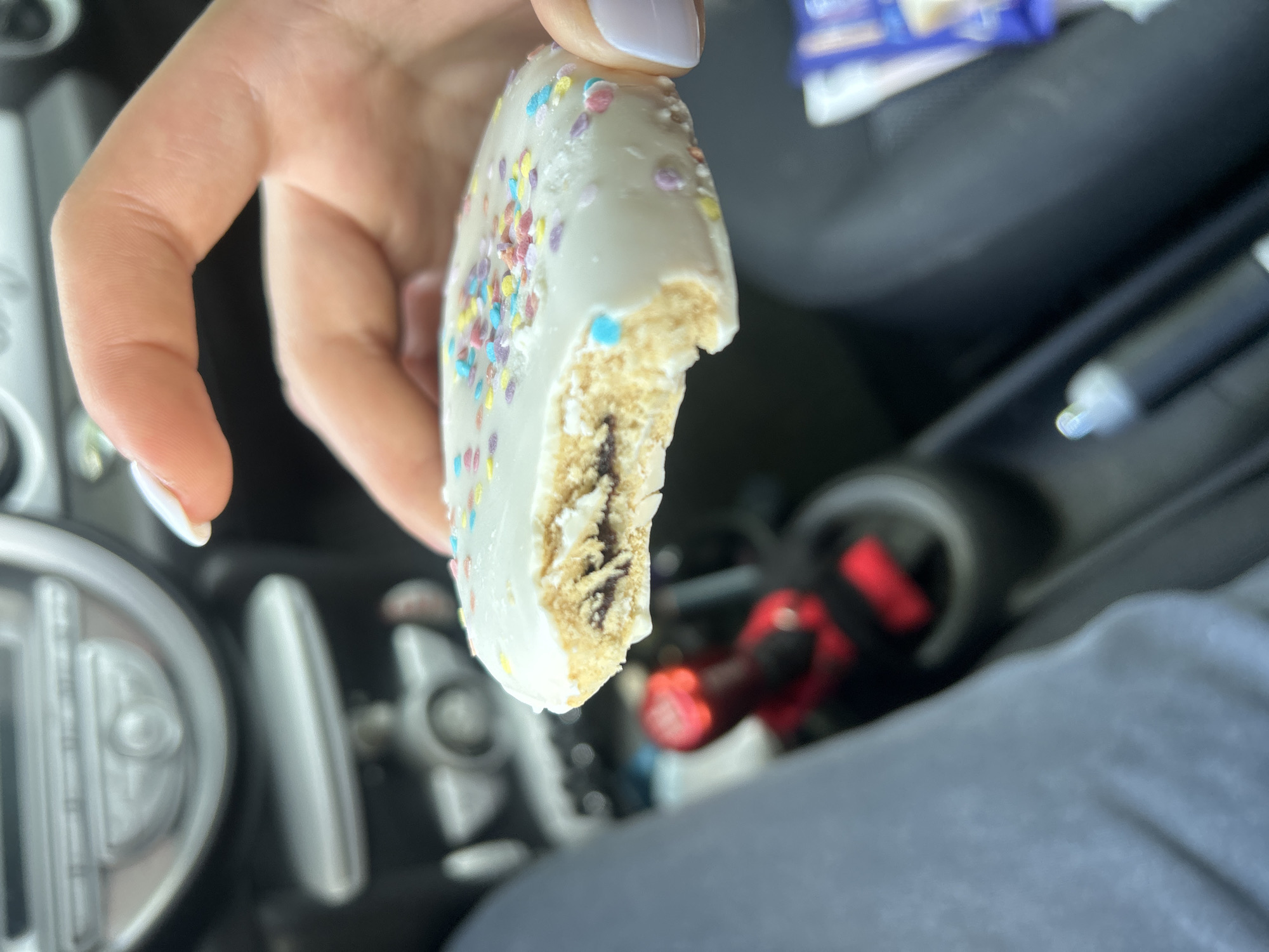 A hand holds a half-eaten cookie with white frosting and colorful sprinkles. The cookie has a visible filling. The setting appears to be inside a car, with elements of the dashboard and seating visible in the background. No evidence suggests that the image was taken in Burgas, Bulgaria.