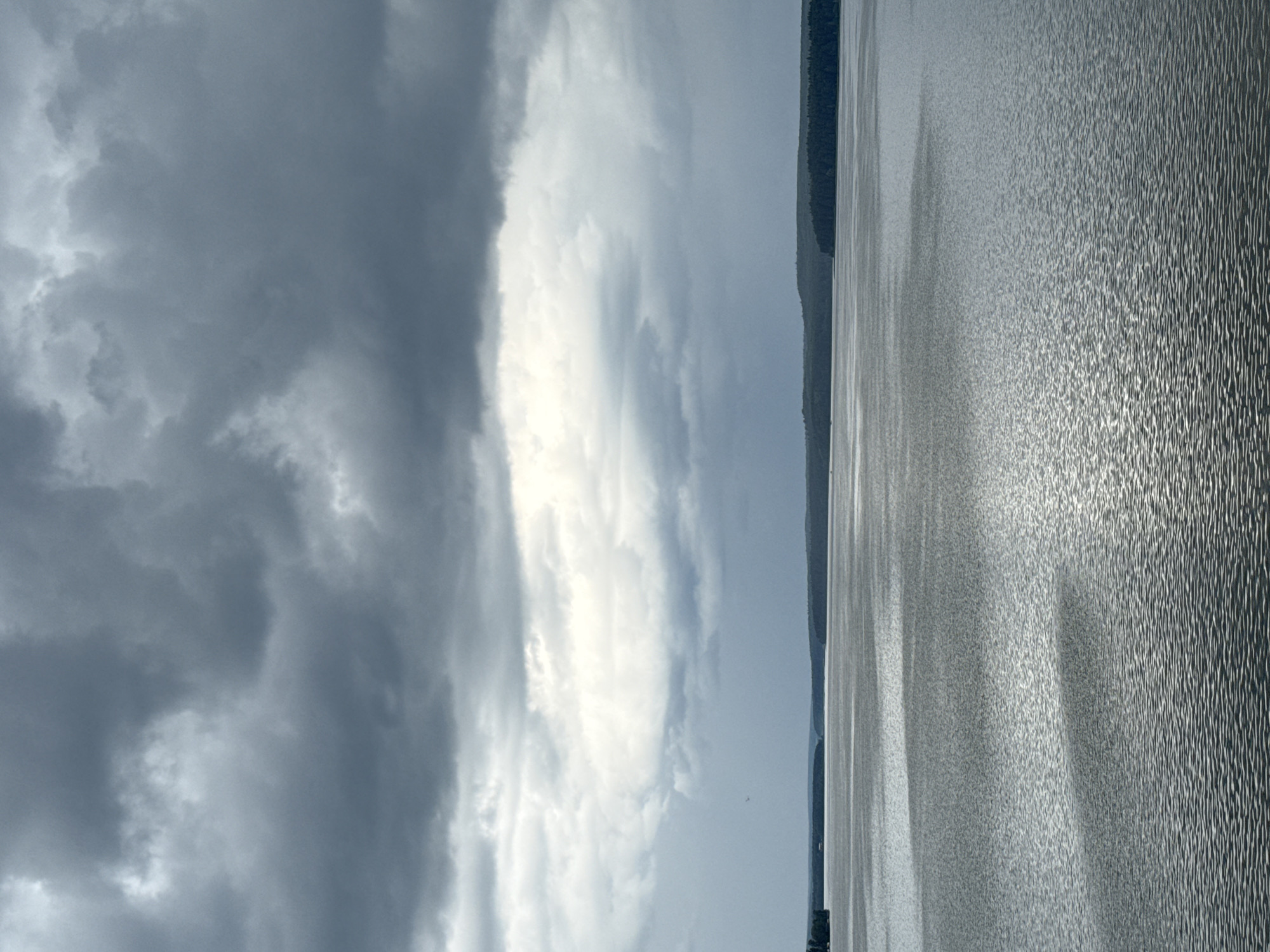 This image depicts a vast expanse of water, likely a lake or large river, under a cloudy sky. The water surface is textured with ripples, catching the light and creating a shimmering effect, particularly on the right side of the frame. A dark, tree-covered shoreline or distant landmass runs along the left edge of the water, creating a stark contrast with the bright reflections.
The sky is dramatic, with dark, heavy clouds dominating the left portion of the image, suggesting impending rain or a recent storm. Towards the center and right, the clouds break, allowing patches of brighter sky and sunlight to filter through, illuminating the water. The overall mood is one of natural grandeur and perhaps a touch of brooding atmosphere due to the heavy clouds.
The scene appears to be a landscape, with no people or specific objects clearly visible. The time of day is difficult to pinpoint precisely, but the quality of light suggests it could be late afternoon or early evening, when the sun is lower in the sky. The location context provided is Votkinsk, Russia, which aligns with the type of expansive water bodies found in that region. There is no visible text in the image.