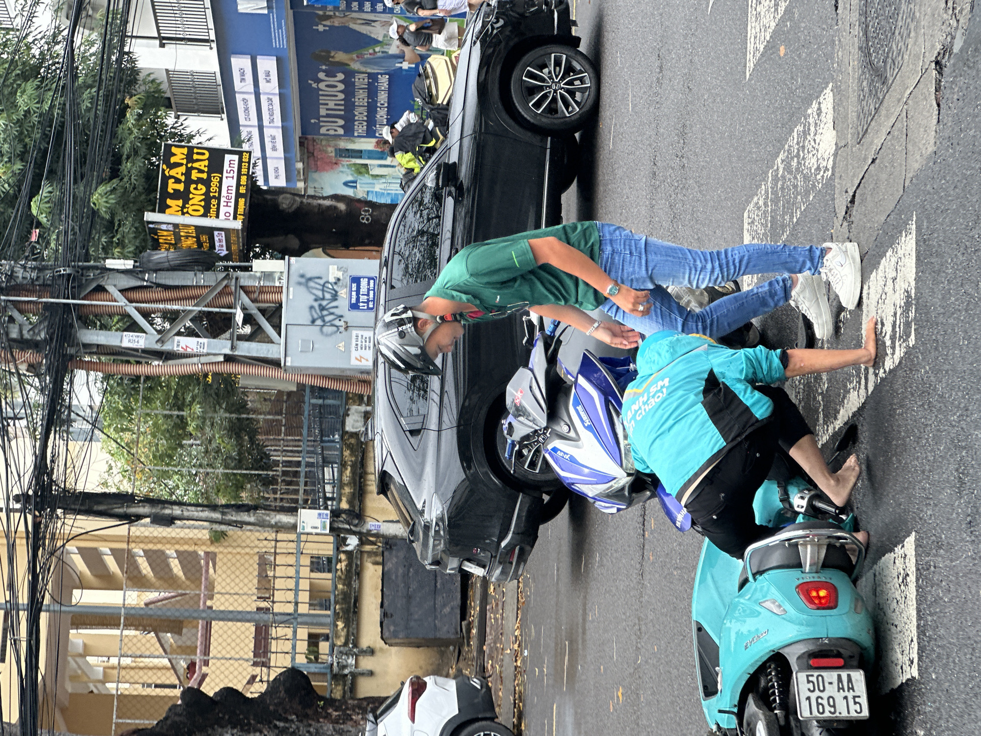 The image captures a street scene in Nha Trang Ward, Vietnam. Two men are interacting near a crosswalk and a black Honda car, which appears to have been in an accident with a teal-colored scooter. One man, wearing a green shirt, jeans, and white sneakers, leans over the scooter. The other man, wearing a turquoise jacket and black shorts, sits barefoot on the scooter. The scooter's license plate reads "50-AA 169.15". In the background, a building with signs is visible, including one that reads "M TẤM ỒNG TÀU (Since 1996)". Power lines are entangled overhead. A white car is partially visible in the lower left corner.