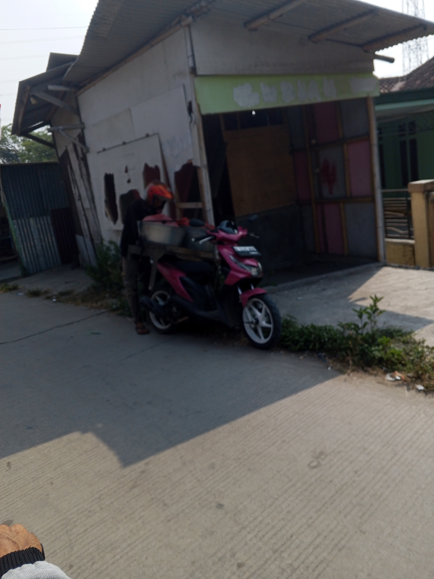 A person wearing an orange helmet and a black shirt stands next to a pink scooter in front of a dilapidated building in Teritih, Indonesia. The building appears to be made of various materials, including corrugated metal and white panels, and has a sloped, weathered roof. The entrance of the building is open, revealing a dark interior with colorful panels on the right side. A metal box or container is on the back of the scooter, and the person is leaning over it. The ground is paved with concrete, and there is vegetation growing along the edge of the road. The scene suggests a rural or semi-urban setting in Indonesia.