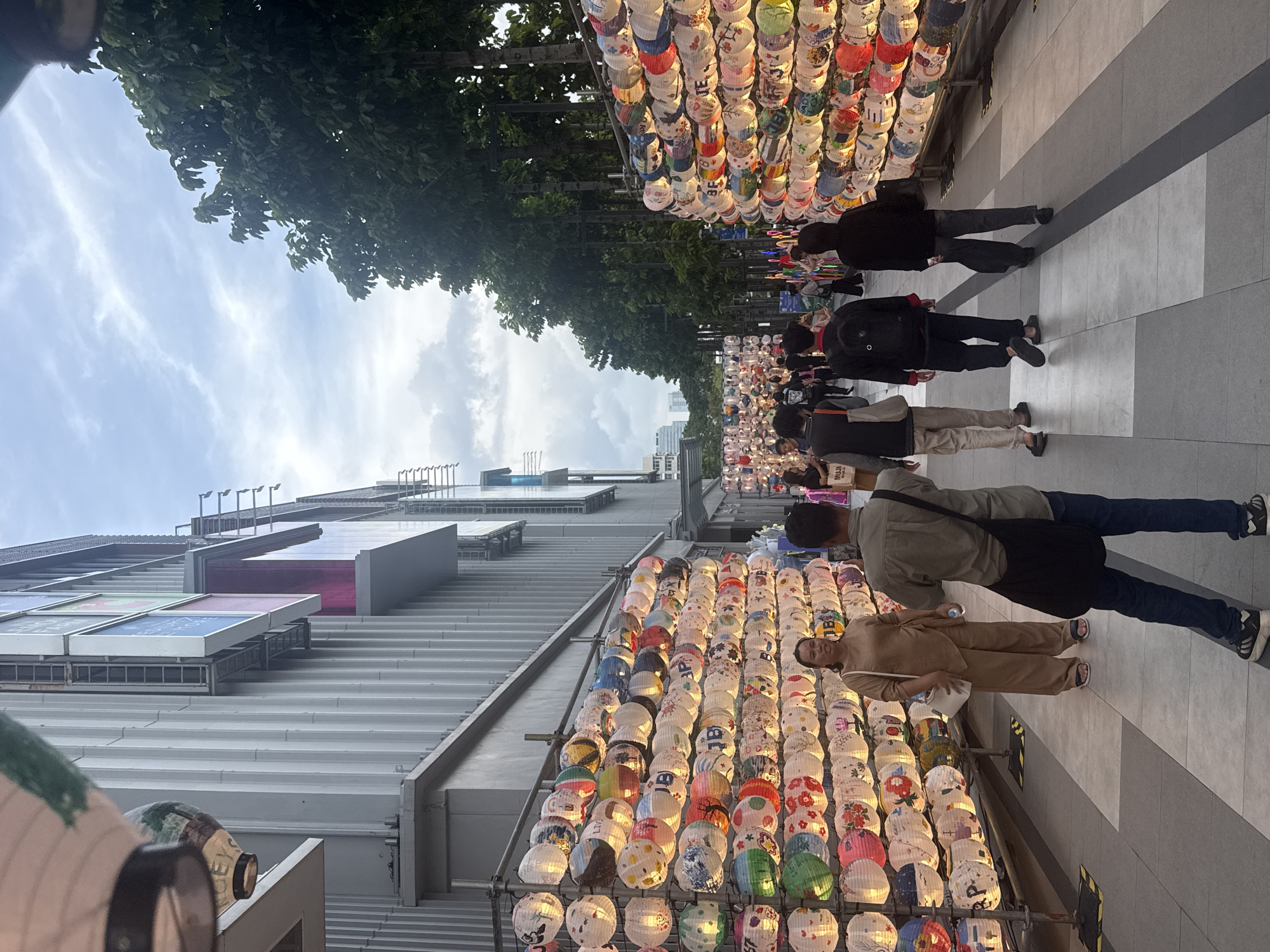 The image depicts a street scene in Bangkok, Thailand, likely during an evening event or festival. The foreground is dominated by rows of colorful, illuminated paper lanterns displayed on metal racks lining both sides of a pedestrian walkway. These lanterns are adorned with various patterns and designs, some featuring Japanese characters.
On the left side of the walkway, a woman in a beige outfit stands looking towards the camera, carrying a white bag. Several people are walking away from the viewer on the walkway, some with backpacks, suggesting they are tourists or shoppers. On the right side, a dense row of trees with dark green foliage forms a natural barrier, with more lanterns visible further down the path. Behind the trees, a modern building complex with a metallic facade and signage is visible, indicating an urban setting. The sky is overcast, with streaks of clouds.
The atmosphere suggests a vibrant public space decorated for a festive occasion. The presence of modern architecture and lush trees alongside the traditional lanterns indicates a blend of contemporary and cultural elements typical of a bustling city like Bangkok.