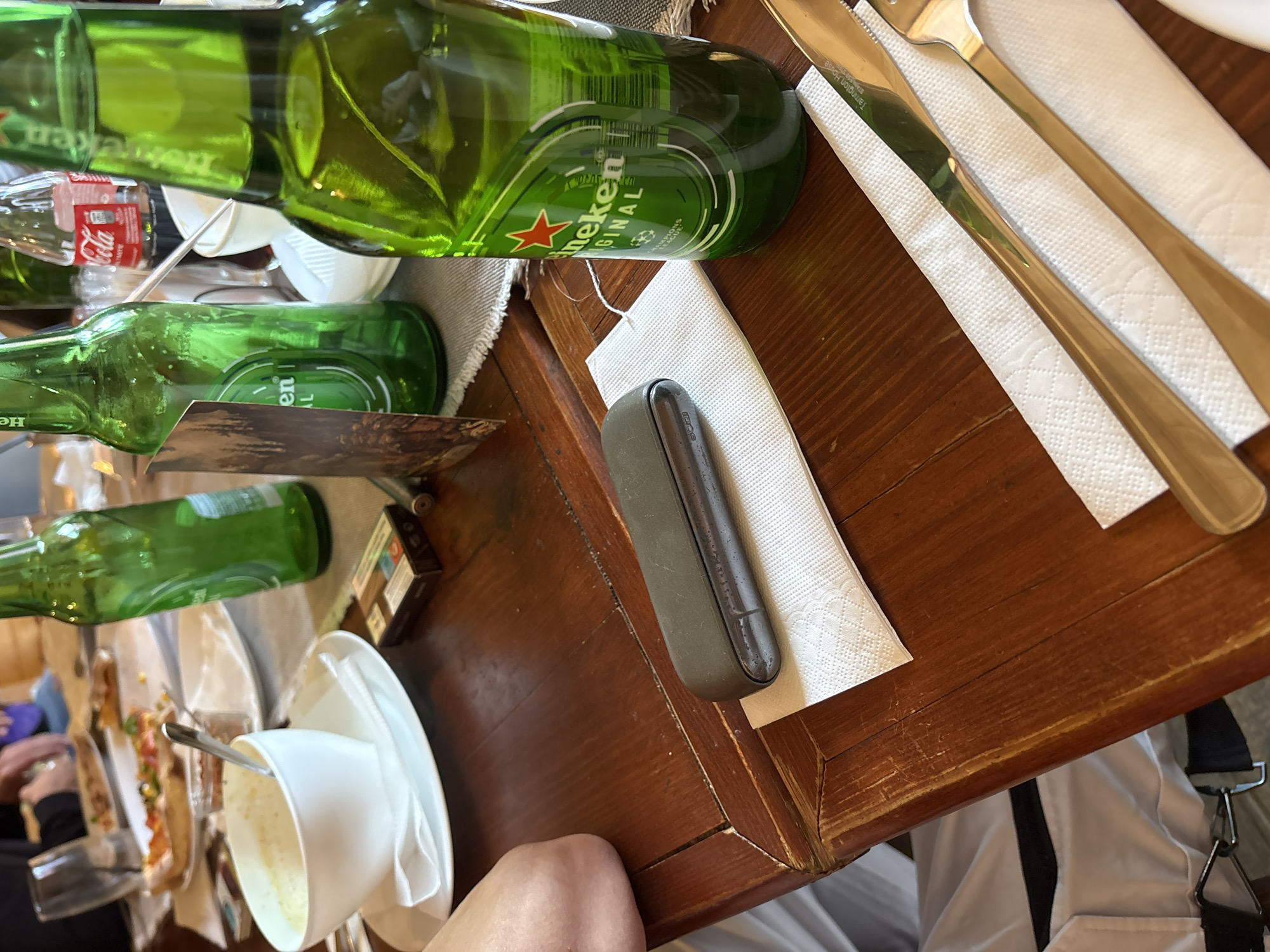A tabletop view features several items, including multiple green beer bottles, a Coca-Cola bottle, a knife and fork on a white napkin, and a dark gray case resting on another white napkin. A white cup and saucer are also visible, along with food items like sandwiches. A person's arm and leg are partially visible in the lower part of the frame. The setting appears to be Ribaritsa, Bulgaria, based on the beer bottles.