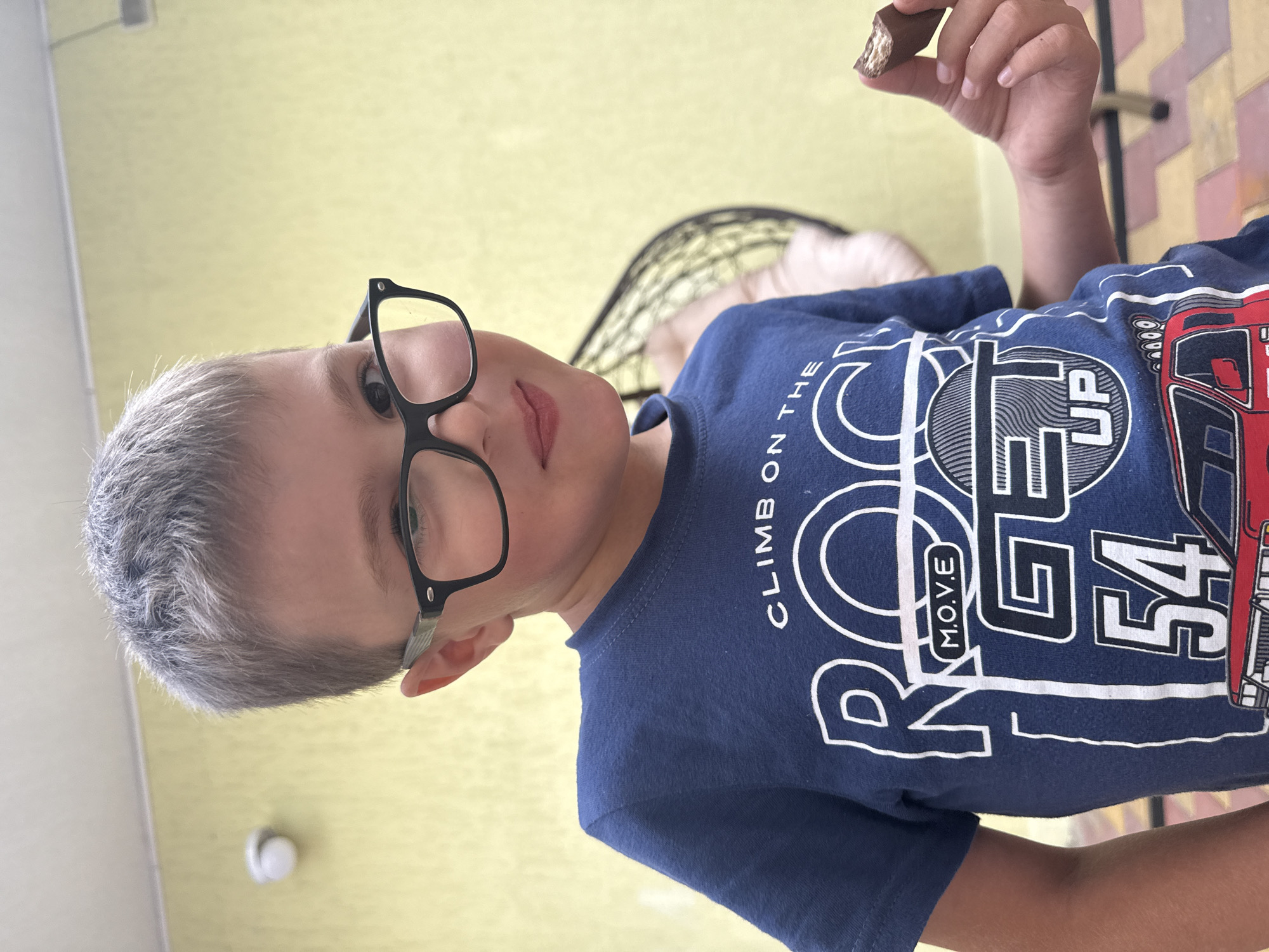 A young boy with short, graying hair and black-rimmed glasses smiles at the camera, holding a bite of a chocolate bar in his right hand. He is wearing a blue t-shirt with white graphic text that reads "CLIMB ON THE ROCK M.O.V.E GET UP 54" and a depiction of a red truck. The background is a textured yellow wall. The image appears to be taken outdoors, possibly in a courtyard or patio area, with cobblestone-like flooring visible at the bottom of the frame. A wicker chair is partially visible in the background. The setting is consistent with a residential or recreational area within the city of Sirius, Russia.