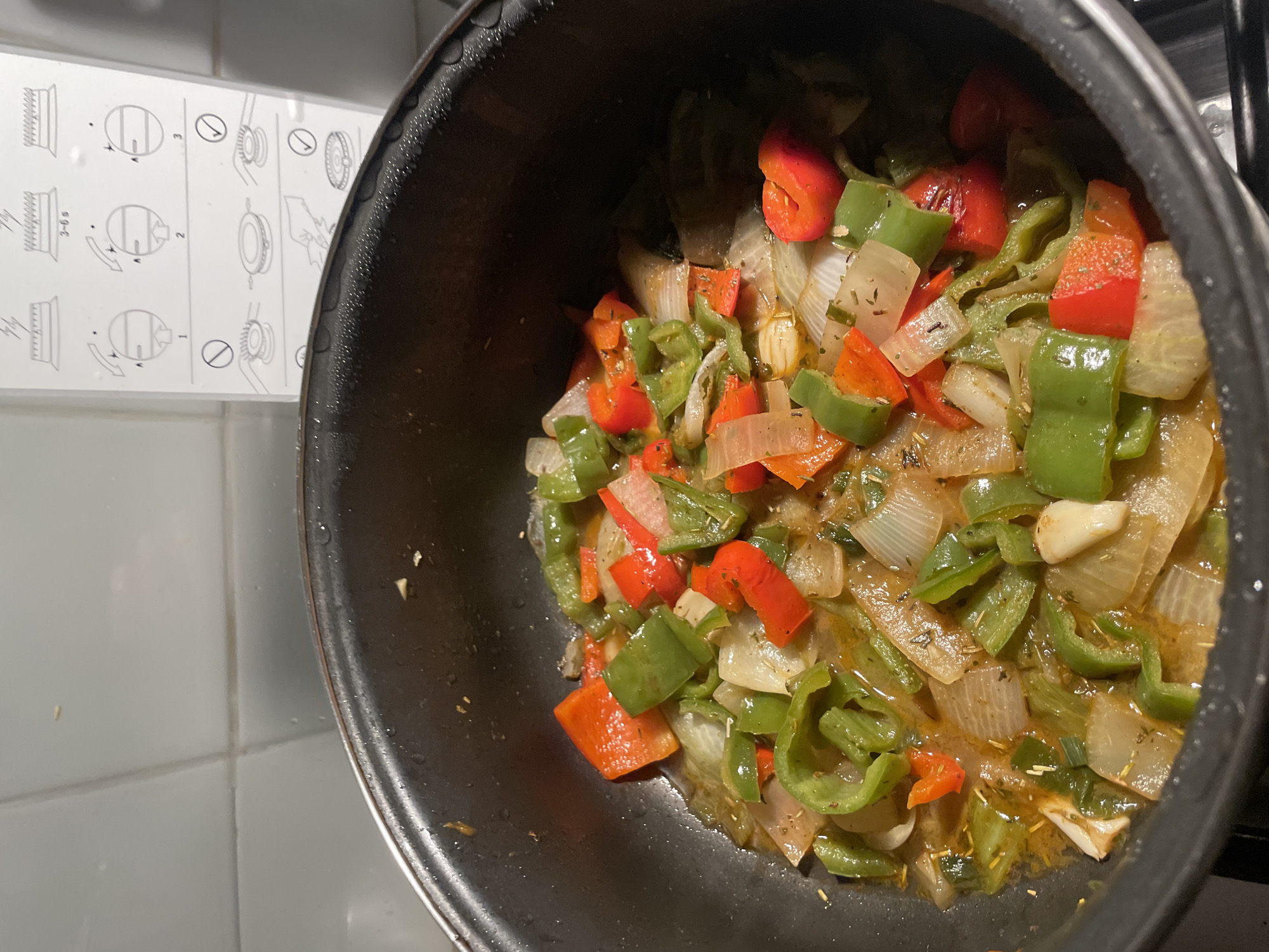 A close-up, high-angle shot shows a dark-colored cooking pan filled with sauteed vegetables, primarily green and red bell peppers and onions. The vegetables appear to be coated in a light, oily sauce. The pan is set on a ceramic tile stovetop next to a white guide with diagrams. This image was taken in an indoor kitchen environment. While it is not possible to determine the precise location, the ceramic tile stovetop and white guide suggest that the photograph may have been taken in Barcelona, Spain.