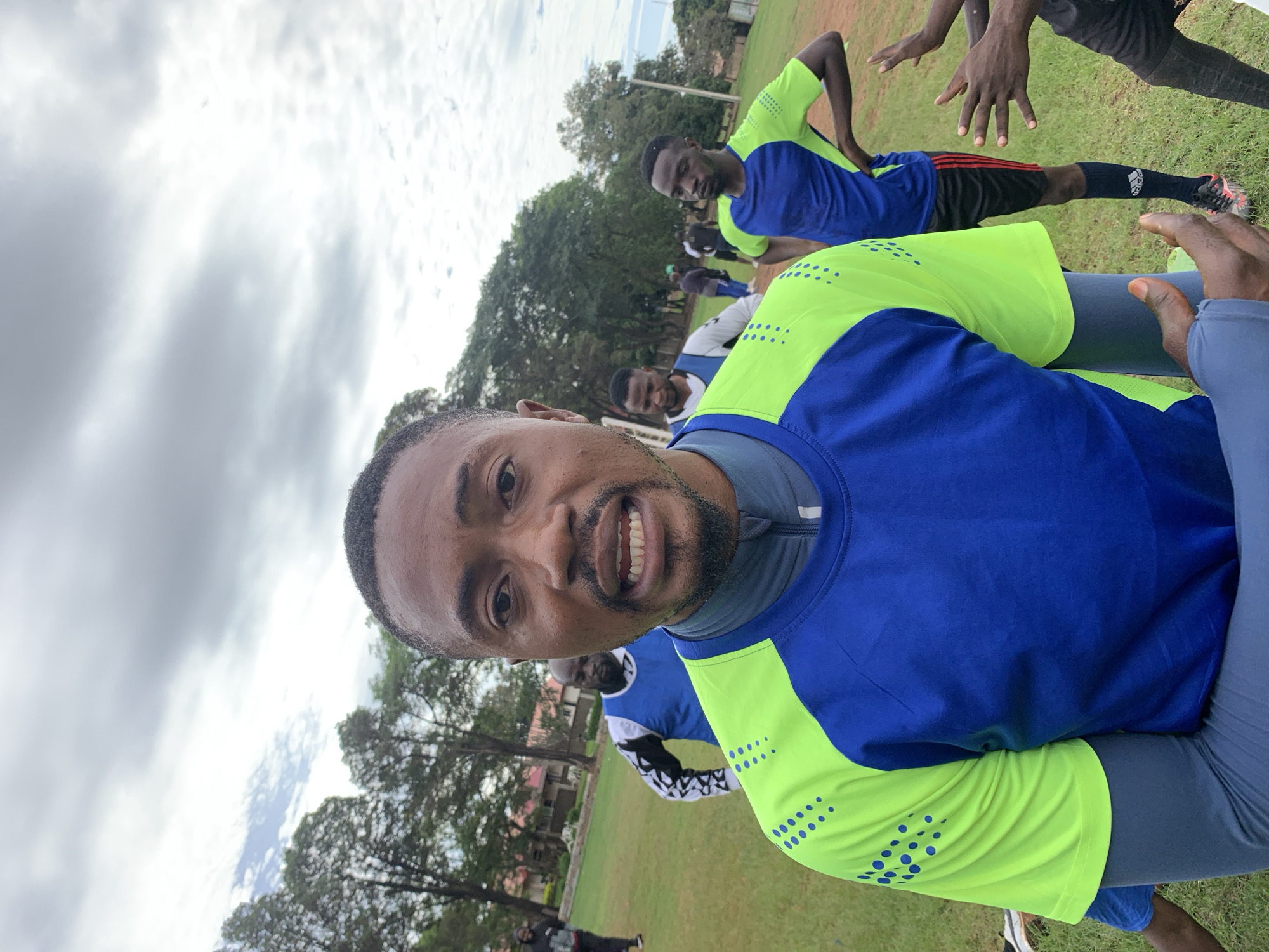 The image captures a close-up of a Black man on a grassy field, presumed to be in Jos, Nigeria, surrounded by other men and trees under an overcast sky. The primary subject, a Black man in his mid-30s with short dark hair, a short beard, and a mustache, faces the camera directly. He is wearing a blue short-sleeved athletic shirt with lime green raglan sleeves that feature a pattern of dark blue dots, over a grey long-sleeved base layer. His mouth is slightly open, revealing his upper teeth.
In the mid-ground, behind and to the left of the main subject, another Black man is visible, wearing a blue training bib over a white long-sleeved shirt with black and white patterned sleeves. To the right of the main subject, a third Black man, also wearing a blue and lime green athletic shirt, black shorts, and dark athletic shoes with red accents, stands with his hands on his hips. Several other individuals are dispersed further in the background, some walking, others standing on the field.
The environment consists of a large expanse of green grass, indicative of a sports field or park, with some areas of brown earth. Numerous tall, leafy trees form a dense green line in the background, and distant buildings with reddish roofs are partially visible beyond the trees. The sky is uniformly grey and overcast.