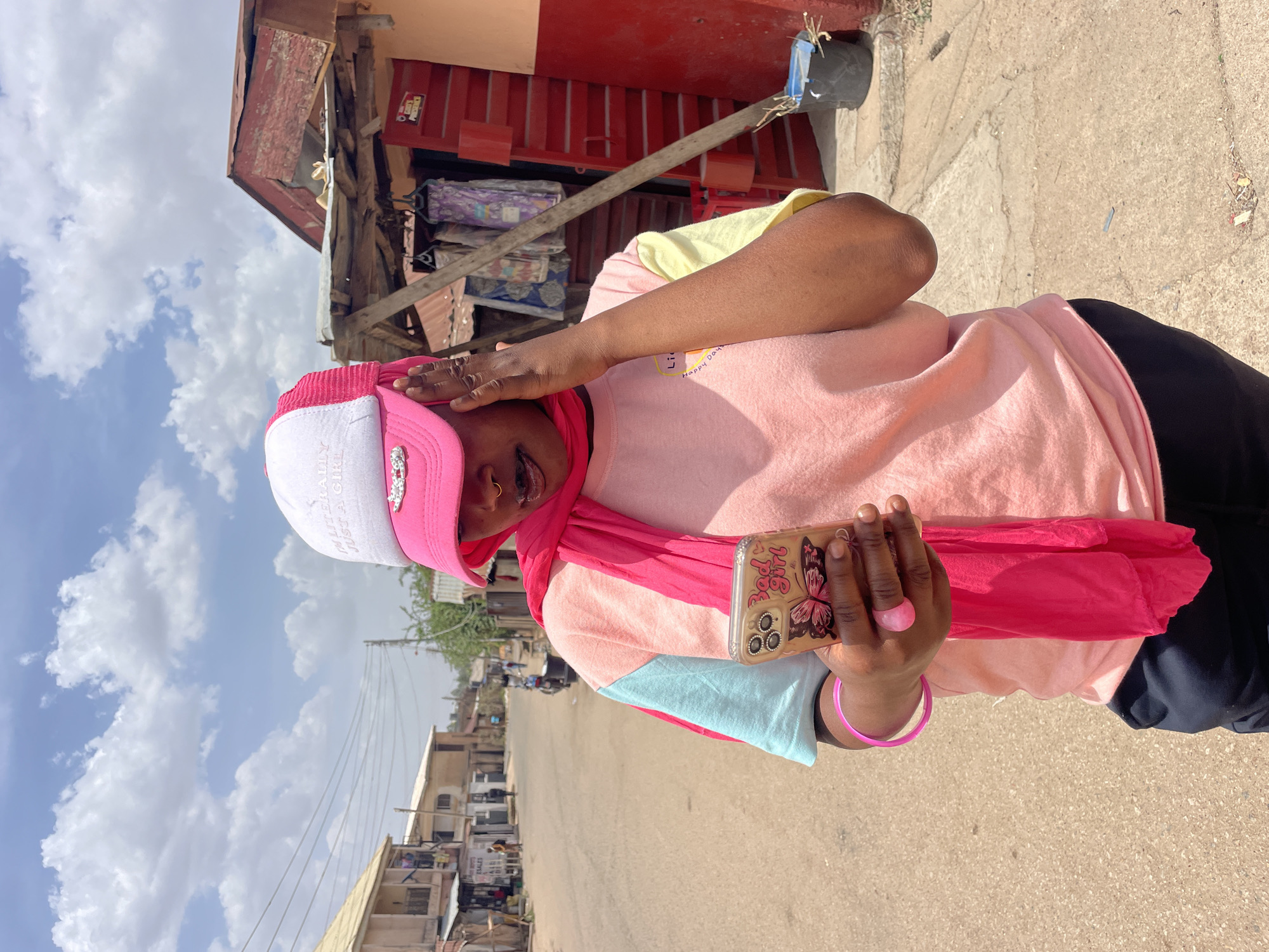A person is depicted outdoors in what appears to be a street scene in Jos, Nigeria, under a bright, sunny sky with scattered white clouds. The image is oriented sideways, but depicts the person standing upright, facing slightly right and looking downwards at a phone held in their left hand. Their right hand is raised to their head, near a pink and white baseball cap.
The individual is wearing a pink and white cap with the text "REALITY GIRL" visible on the white section. A vibrant pink scarf is draped around their neck and partially covers their head. They are dressed in a color-block t-shirt featuring shades of pink, yellow, and light blue, with the words "LITTLE HAPPY DAYS" faintly visible on the front. Black trousers complete their attire. Accessories include a small nose ring, a pink ring on their right index finger, and a pink bangle on their left wrist. The phone case features a prominent butterfly design and the text "Bad Girl".
The background includes various rustic buildings with corrugated iron roofs and some red-painted sections. One building has a red sign with "Prestige" visible. Further down the street, another building shows a sign with "MOTO SALES". The ground is a light-colored, unpaved or concrete surface, suggesting a typical street or pathway in an urban or semi-urban area. The strong sunlight indicates it is daytime.