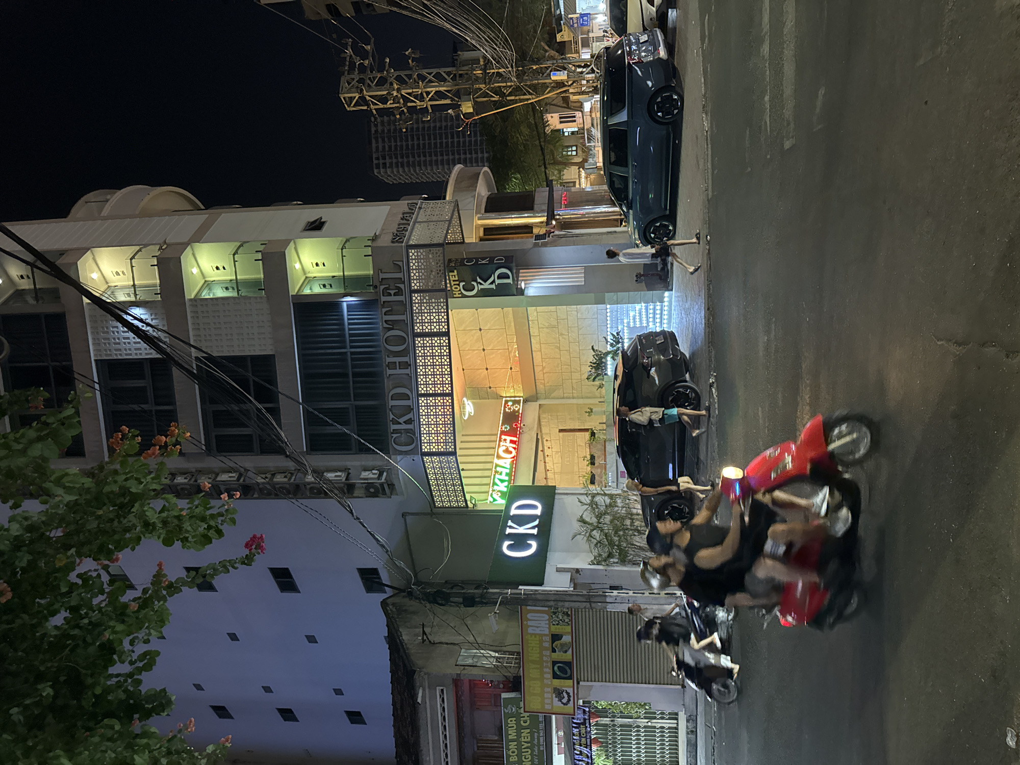 This media file depicts a night scene on a street in Nha Trang, Vietnam, likely in an urban area given the architecture and overhead power lines. The primary subjects are people and vehicles on the street, suggesting a typical evening atmosphere.
Several individuals are visible, some on a red scooter and others standing near parked cars. The scooter appears to be in motion, with a driver and a passenger, indicating people are traveling or commuting. The presence of parked cars, including a dark-colored sedan, suggests a commercial or residential area where parking is available.
The setting is characterized by buildings with illuminated signs, notably those for "CKD HOTEL" and "CKD HOME". Another sign reads "V KHACH", which likely translates to "Guest" in Vietnamese, further confirming the presence of a hotel. Other signs with Vietnamese text, such as "BỐN MÙA NGUYÊN CHẤ" and "ĐỒ GỖ MỸ NGHỆ BẢO", indicate various businesses operating in the vicinity, possibly shops or service providers. The time of day is clearly night, as evidenced by the dark sky and the artificial lighting from the signs and buildings. There are no obvious cues about the weather, but the scene appears dry.
Notable details include the vibrant green of the "CKD" sign, the red color of the scooter, and the architectural style of the hotel building with its patterned facade. The overall impression is that of a lively street at night, with commerce and daily life continuing.