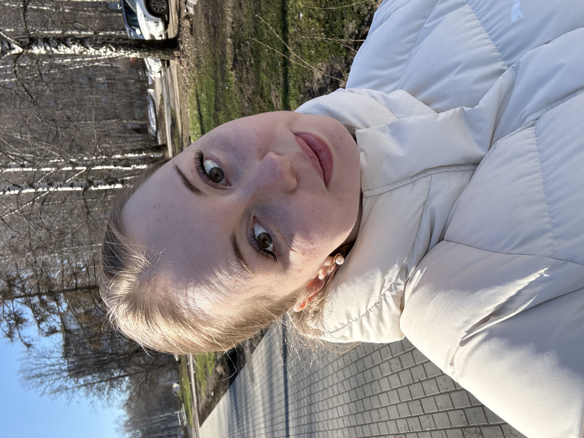 A woman is seen in a close-up, angled selfie outdoors. She is wearing a white puffer jacket and a pearl earring is visible. The background consists of trees without leaves and patches of green grass, suggesting a park or a green area in a city. A gray brick sidewalk is visible in the bottom right corner of the frame. A vehicle, likely a car, is partly visible in the background, suggesting a street or parking area nearby.