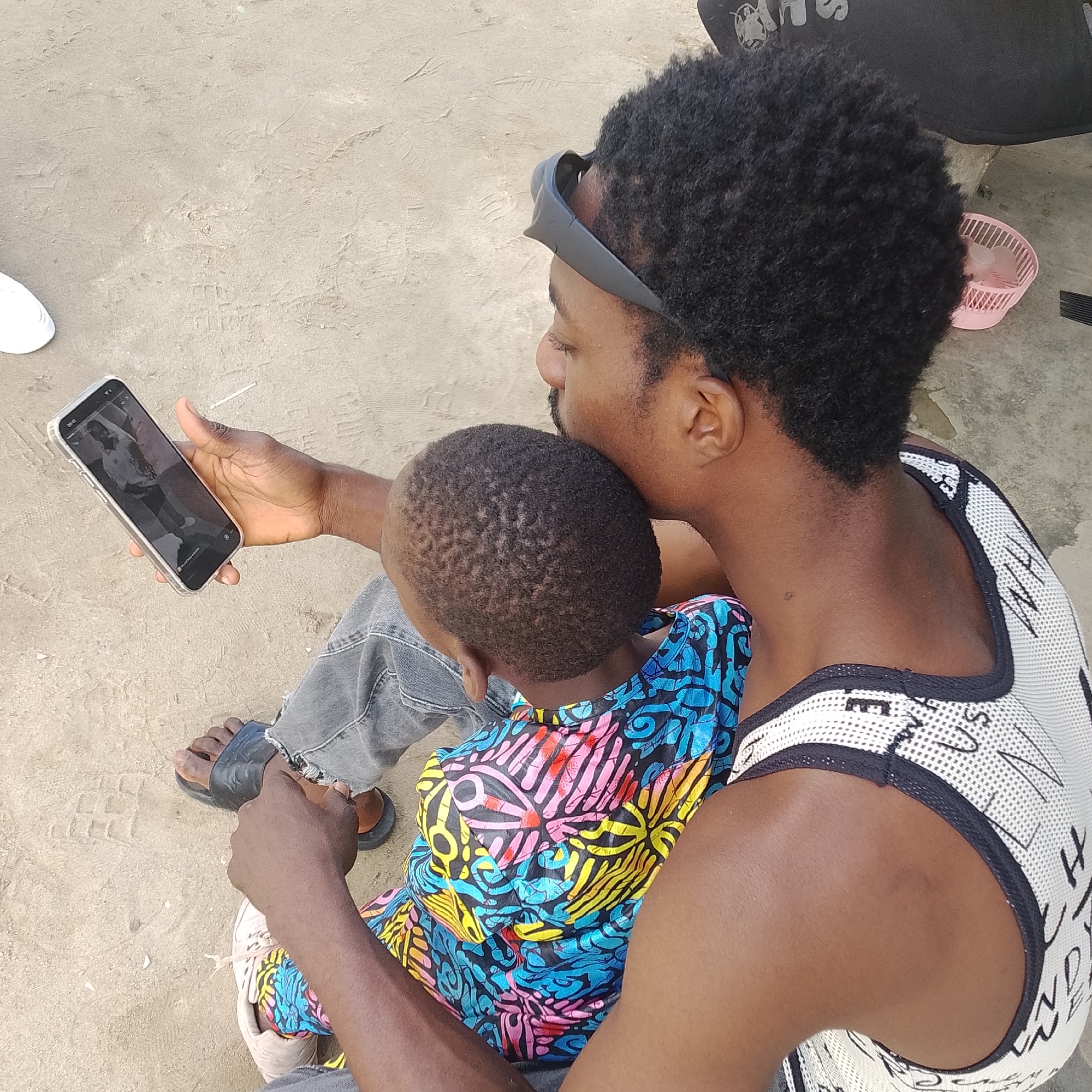 In Ado, Nigeria, a man wearing a white tank top with black trim and text, along with denim jeans and black sandals, is seated on the ground with a young child. The child is wearing a colorful, patterned garment. The man is holding a smartphone, displaying an image, and appears to be showing it to the child. He has sunglasses perched on his head. The ground is sandy and appears to be an outdoor location. A pink basket with what appear to be plastic cups and utensils is visible to the right of the man. The overall scene depicts a moment of interaction between the man and child in a casual outdoor setting.