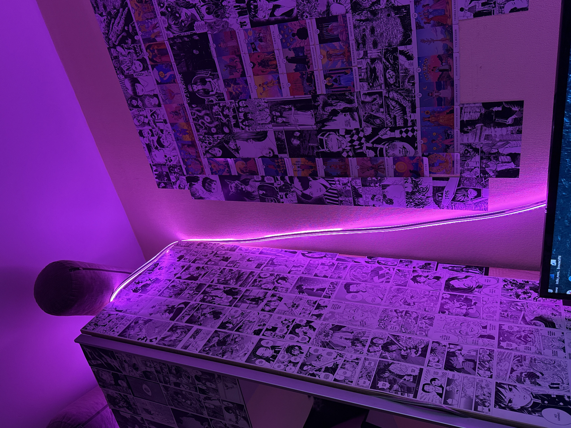 This image depicts a room, likely a personal study or bedroom, illuminated by intense purple LED lighting, situated in Moscow, Russia. The dominant features are two large collages of printed media adhered to vertical surfaces.
On the left, a tall, narrow structure, possibly a cabinet or wardrobe, is entirely covered with numerous black-and-white manga panels, primarily from the *Demon Slayer: Kimetsu no Yaiba* series, featuring characters such as Giyu Tomioka and Shinobu Kocho. A bright purple LED strip runs along the top edge of this structure and continues vertically down the adjacent wall.
The right wall features an extensive collage of diverse prints, arranged in a grid-like fashion. This collage includes a mixture of black-and-white manga illustrations, some appearing to be in the style of horror manga (e.g., Junji Ito), alongside numerous colored tarot card designs. These tarot cards are labeled with their names in Russian Cyrillic, such as "БАШНЯ" (Tower), "ШУТ" (Fool), "СМЕРТЬ" (Death), "ЛУНА" (Moon), "КОЛЕСО ФОРТУНЫ" (Wheel of Fortune), and "ВЕРХОВНАЯ ЖРИЦА" (High Priestess), among many others, including "ГРОБ" (Coffin), a Lenormand card. Some additional prints include stylized character art and text like "ХУДОЖНИК" (Artist).
Above the left structure, a cylindrical, fabric-covered object, potentially a lampshade or speaker, hangs from the ceiling via a cable, also bathed in the purple light. At the bottom edge of the image, the upper portion of a computer monitor is visible, displaying a dark interface with white text including "lodka_more... BrowseVPN", indicating a virtual private network service. No individuals are present in the scene.