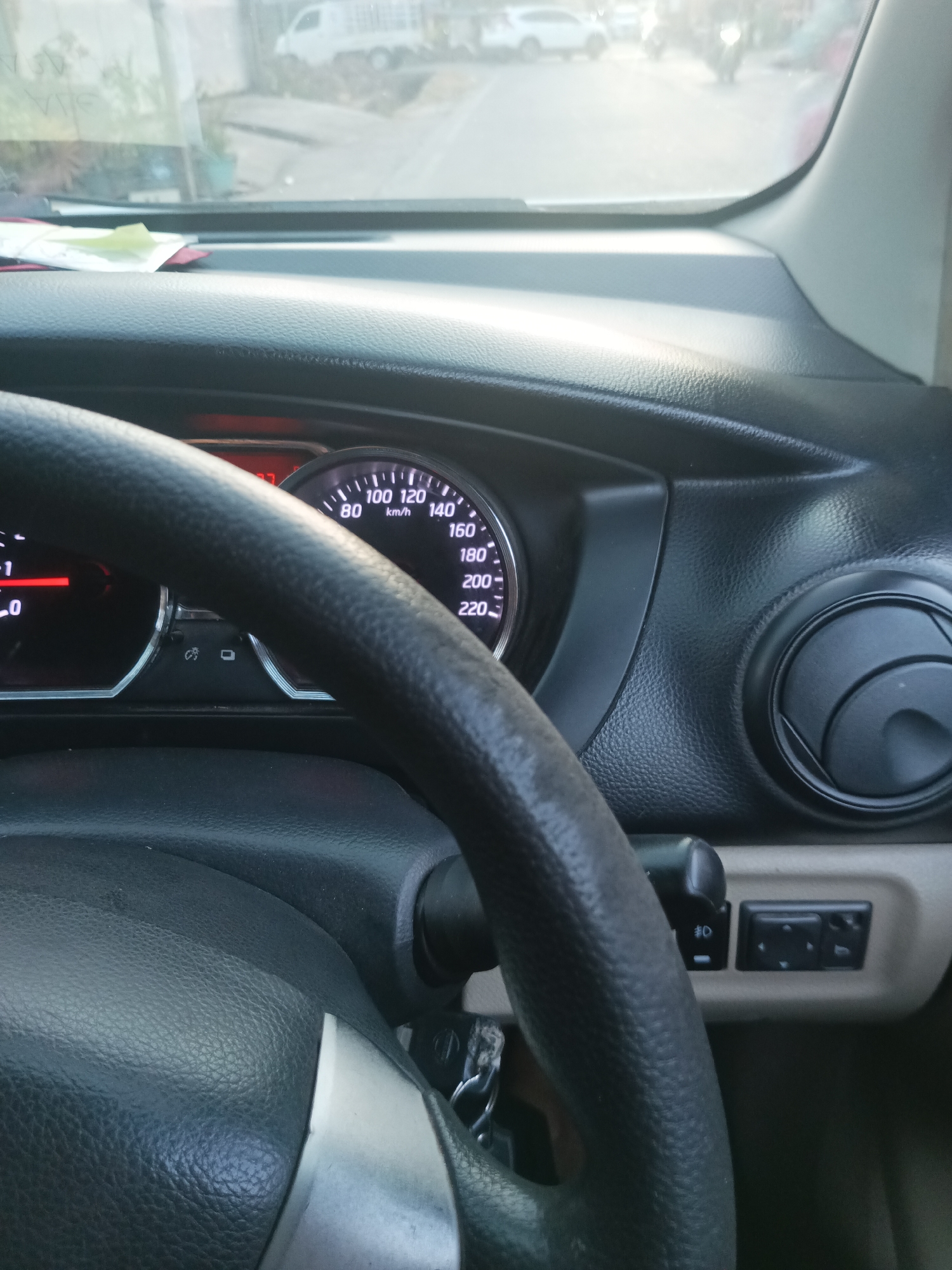 The image shows the interior of a car, likely a Nissan Grand Livina based on the dashboard design, looking towards the front windshield and the road outside. A black steering wheel is prominent in the foreground, partially obscuring the car's instrument cluster which displays a speedometer with a needle indicating around 100 km/h, and what appears to be a fuel gauge with a reading of 0. To the right of the speedometer is a circular air vent. Below the instrument cluster, there are two small buttons, one with a symbol that looks like a fog light. Through the windshield, a blurred street scene is visible with a white pickup truck and other vehicles, suggesting the car is either parked or moving slowly on a road in Pangkajene, Indonesia. The lighting indicates it is daytime.