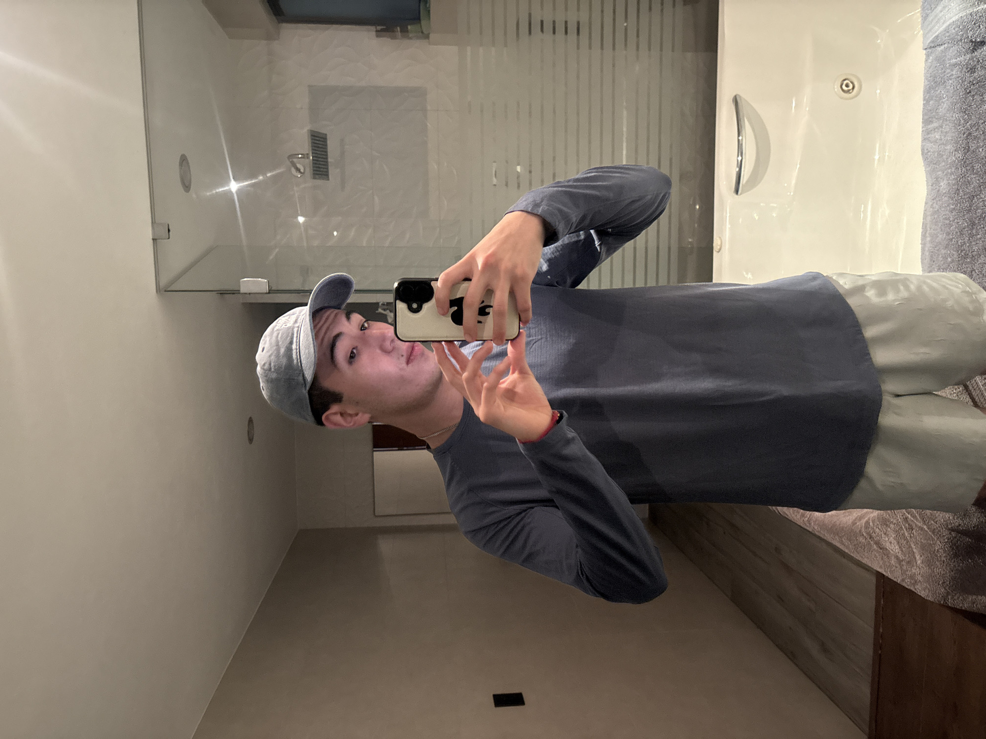 A young man wearing a light blue baseball cap and a grey long-sleeved shirt is taking a selfie in a bathroom. He's holding a phone with a light-colored case and making a hand gesture. The bathroom features a glass-enclosed shower, white walls, a white bathtub with a silver handle and nozzle, and a grey towel hanging near the tub. There is a wooden floor or cabinet beneath the towel and a dark rectangular object is on the floor. The location is likely in San Bernardino Tlaxcalancingo, Mexico.
