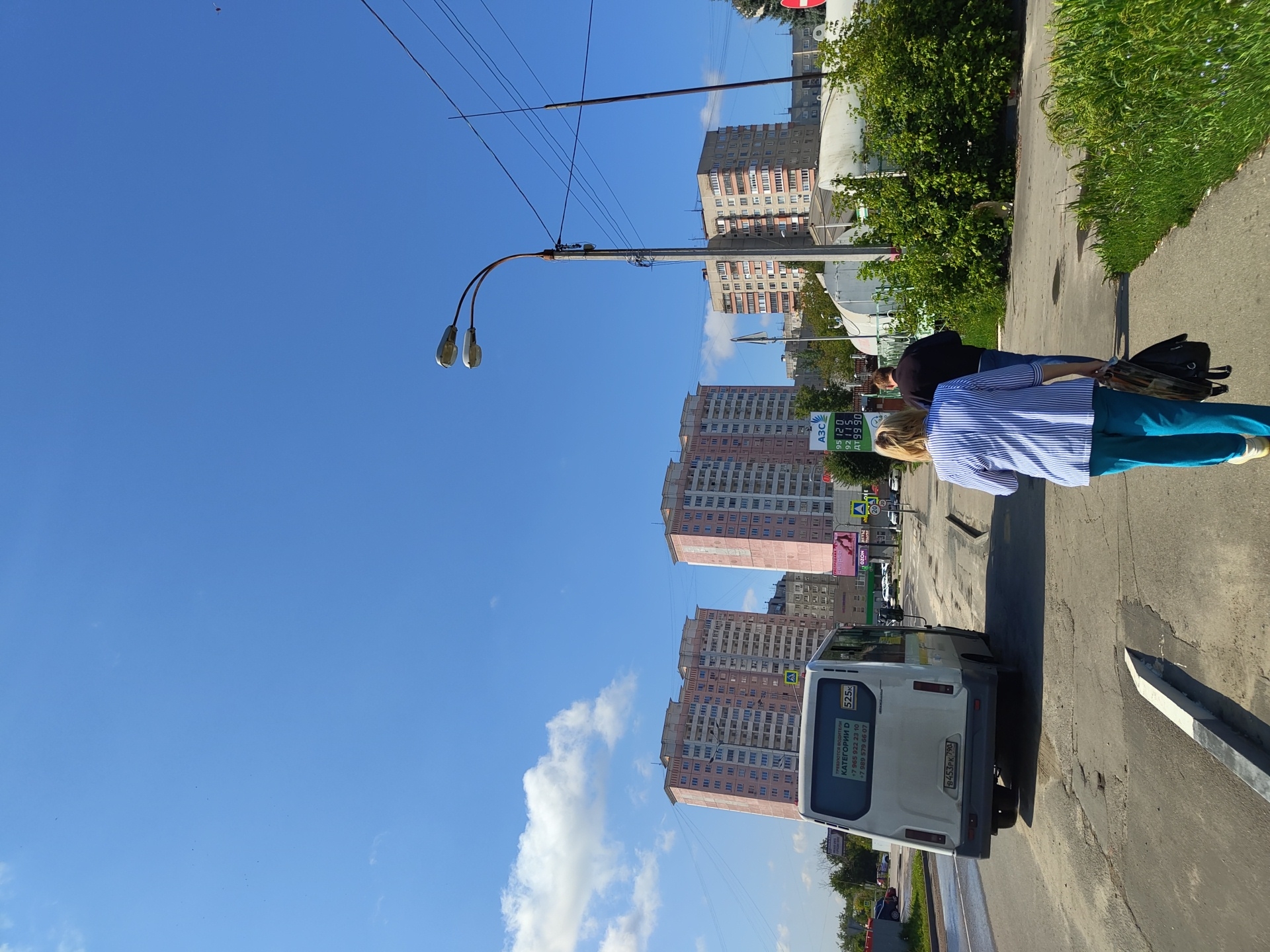 The image depicts a street scene in Zhukovsky, Russia, on a clear, sunny day with a few scattered clouds. In the foreground, two individuals are walking away from the camera on a sidewalk. The person on the left is a woman wearing a blue and white striped shirt and blue pants, carrying a black bag. The person on the right is a man wearing a dark shirt and blue jeans.
A white minibus is parked on the left side of the street, facing away from the camera. It has a license plate that reads "B453PK 790". On the rear window of the minibus, there is text indicating "КАTEГОРИИ D" and phone numbers.
Further down the street, there are several tall, multi-story apartment buildings with pink facades. One building on the left has text and signage visible, including what appears to be a gas station price display with "АЗС" and fuel prices. A streetlamp with two lights stands on the right side of the road, with power lines running overhead. In the background, there are more buildings and trees, and a large white tent-like structure. The road surface appears to be asphalt with some visible wear and tear. The overall environment suggests an urban residential or commercial area.