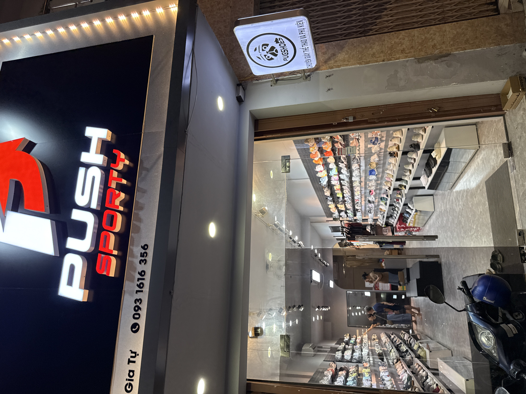 This is an eye-level, medium shot of the exterior of a shoe store. The store's name, "PUSH SPORTY," is displayed prominently on a large, illuminated sign above the entrance. Below the store name, the phone number "093 1616 356" is visible. The store appears to be located in Nha Trang, Vietnam, as indicated by the text "Gia Tự" on the signage.
The store has a large glass entrance, through which the interior is visible. Inside, shelves are stocked with numerous pairs of athletic shoes. Two customers, a man and a woman, are browsing the merchandise. A motorcycle is parked in front of the store. To the right of the entrance, a square sign with a cartoon pug face and the text "PUGSHOES" is mounted on the wall, along with Vietnamese text that translates to "Sports Shoes and Accessories." The street outside is paved, and there's a metal security grate to the right of the store. The scene is lit by a combination of the store's internal lighting and external streetlights.