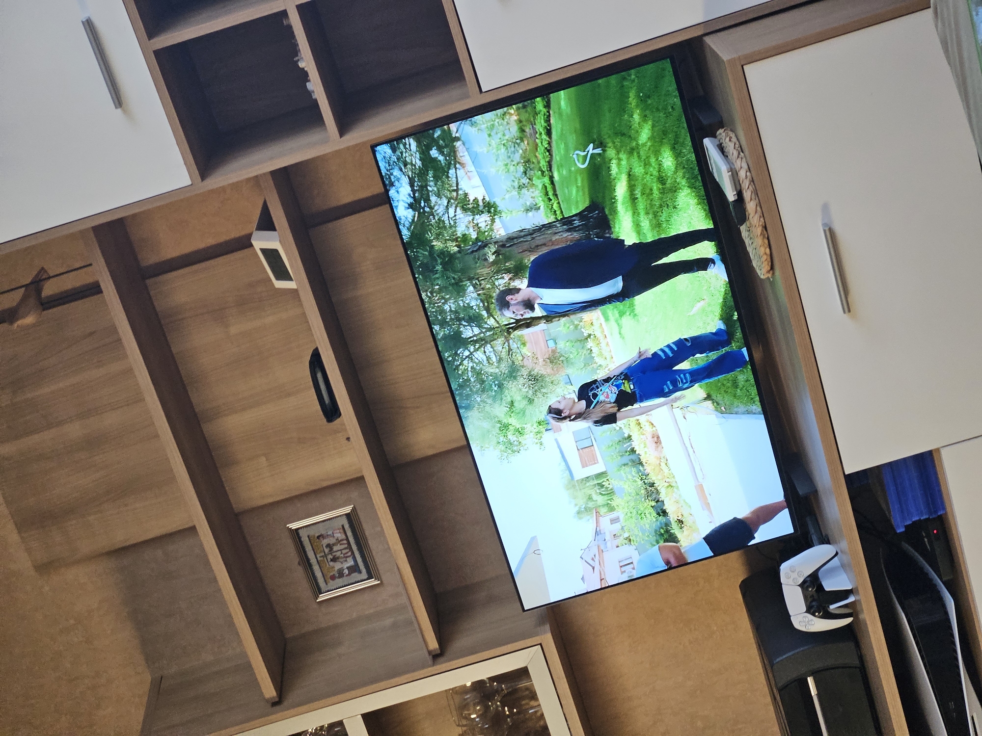 An indoor shot captures a television screen displaying a scene with two individuals in a grassy outdoor setting. The television is positioned on a wooden shelf unit. To the left of the television, a black game console and a white game controller are visible on another shelf. Above the television, on a higher shelf, a framed picture and a small black object are present. The background consists of a wall with a wood-grain finish and part of a white cabinet. The room appears to be a living area, possibly a media room. The image is slightly tilted, suggesting it was taken at an angle.