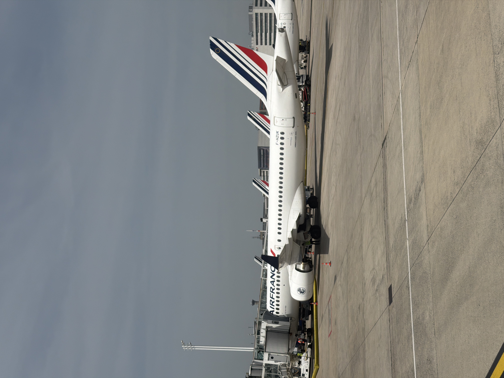 The image shows a row of Air France airplanes parked at an airport apron. The planes are white with the Air France logo on the side and the French flag on the tail. The location is Le Mesnil-Amelot, France, given the presence of Air France planes, which are based at Charles de Gaulle Airport near Le Mesnil-Amelot. There are airport ground support equipment, including baggage handling vehicles and ground power units, clustered around the airplanes, suggesting they are either preparing for departure or have recently arrived. There are shadows indicating daylight. The sky is overcast. The apron itself is a concrete surface with painted taxiway lines.