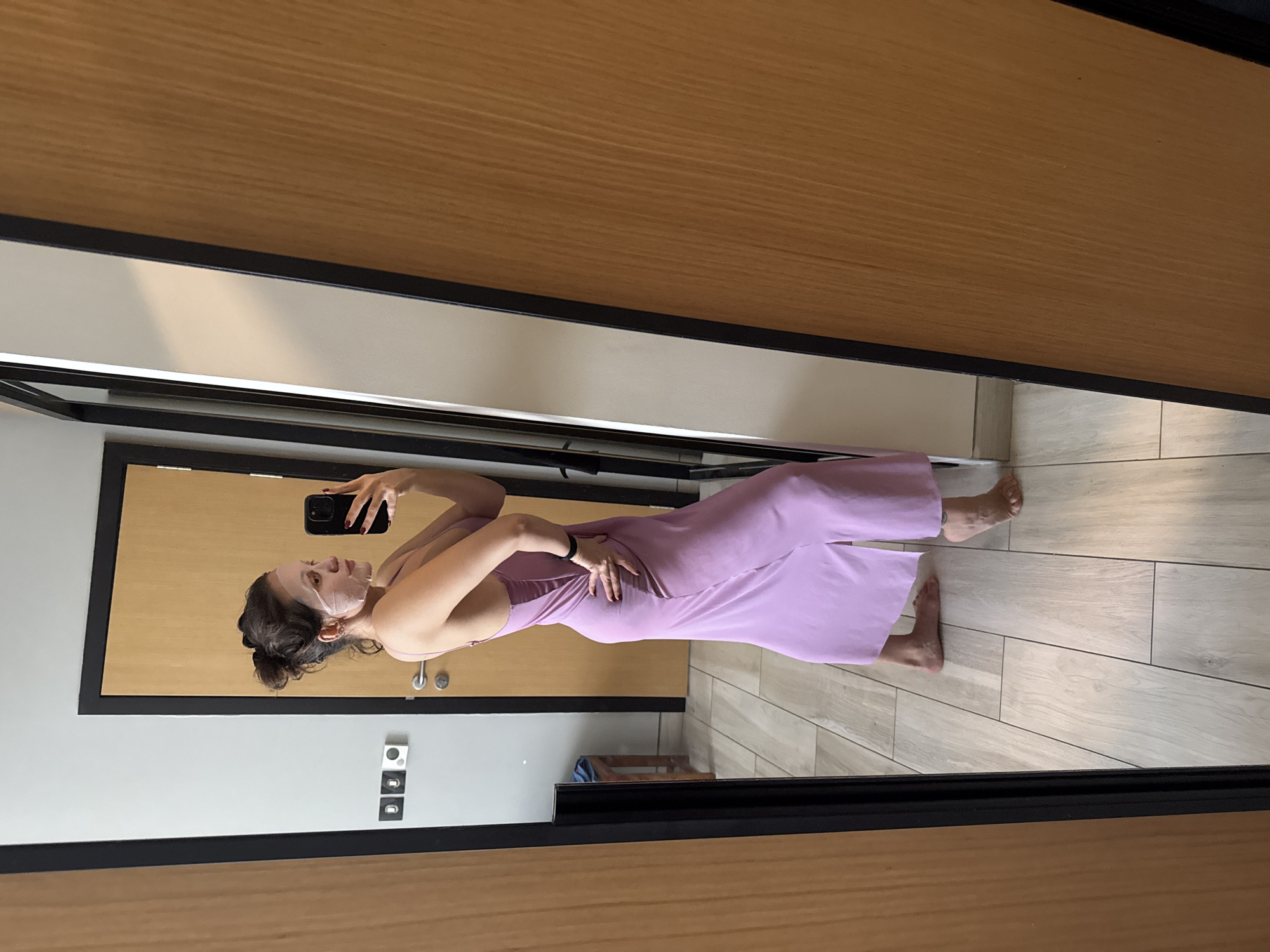 A woman wearing a lavender jumpsuit and a facial mask is taking a selfie in front of a mirror. The woman has her hair pulled back in a bun. The background shows a gray wall with electrical outlets and a wooden door. The flooring is light-colored tile. The location appears to be an interior space, possibly a hotel room. There is no explicit evidence that indicates this location is in Dubai, United Arab Emirates.