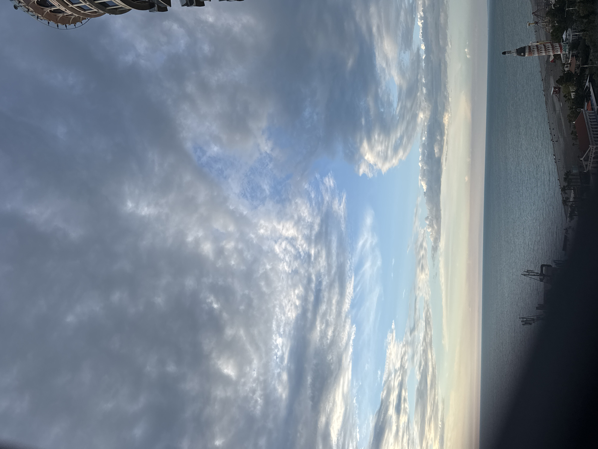 The image shows a coastal view in Batumi, Georgia. The Black Sea stretches along the right side of the frame, with the shoreline visible. Along the shore, buildings and structures can be identified. Prominently, a tower-like building with distinct architectural elements stands out. The sky, taking up the majority of the view, is filled with various cloud formations, ranging from dark gray to light, wispy clouds, with patches of blue sky visible between them. The overall scene suggests an early morning or late afternoon lighting with soft colors. Part of a building can be seen in the top-left corner of the frame.