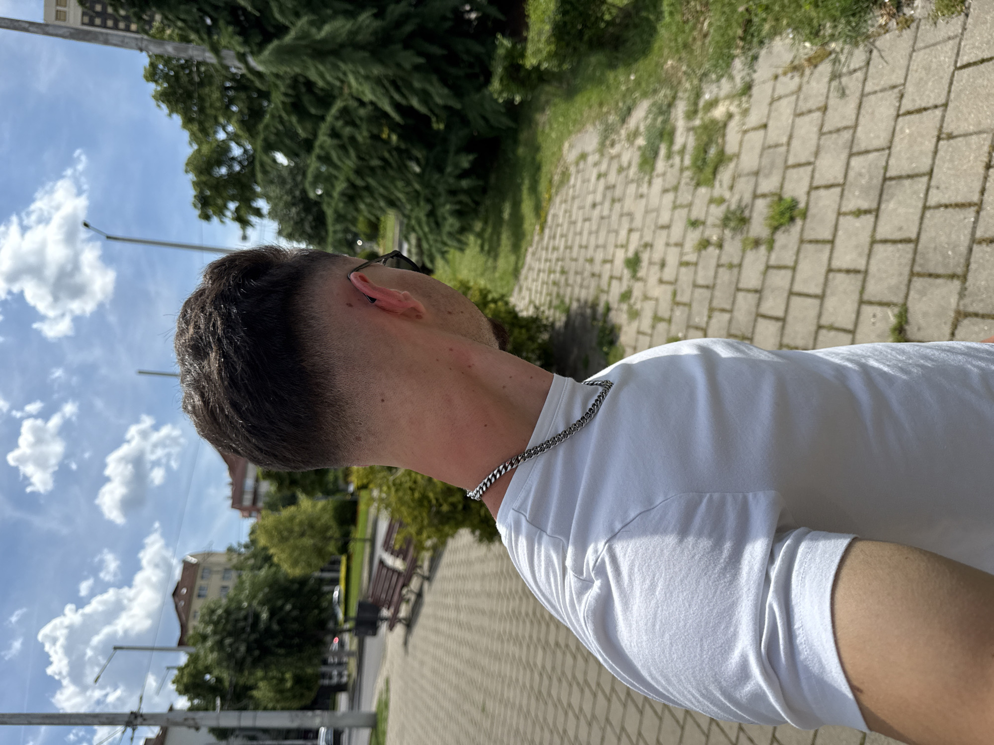 A person is walking outdoors, seen from the shoulder up, with their back to the camera. They are wearing a white t-shirt and a silver chain necklace. The person is walking on a paved sidewalk lined with greenery. In the background, there are buildings, trees, benches, and a street with cars. The sky is blue with white clouds. The image is taken on a sunny day.