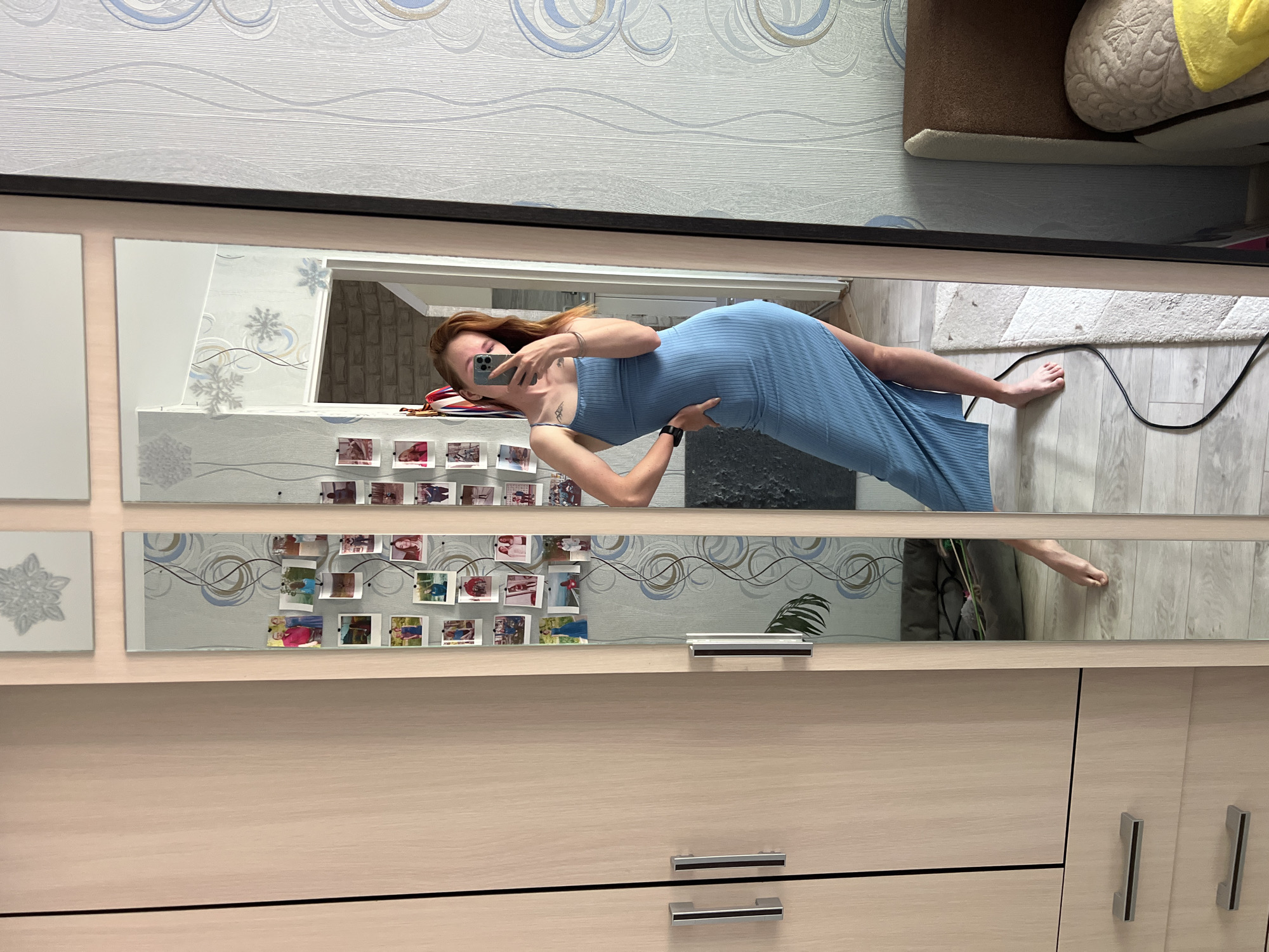 A young woman is seen in a full-length mirror attached to a piece of furniture, possibly a wardrobe or dresser. She is wearing a light blue, form-fitting dress and holding a phone, seemingly taking a selfie. The woman is posing with her left hand on her hip.
The reflection reveals details of the interior of a room. The wall behind the woman is decorated with patterned wallpaper. There are photos and decorative elements placed on a shelf or ledge below the mirror. In the background, there's a glimpse of a floor, a rug, and possibly a couch or cushioned object.
The location is indicated as Chaikovsky, Russia, based on image data not visible.