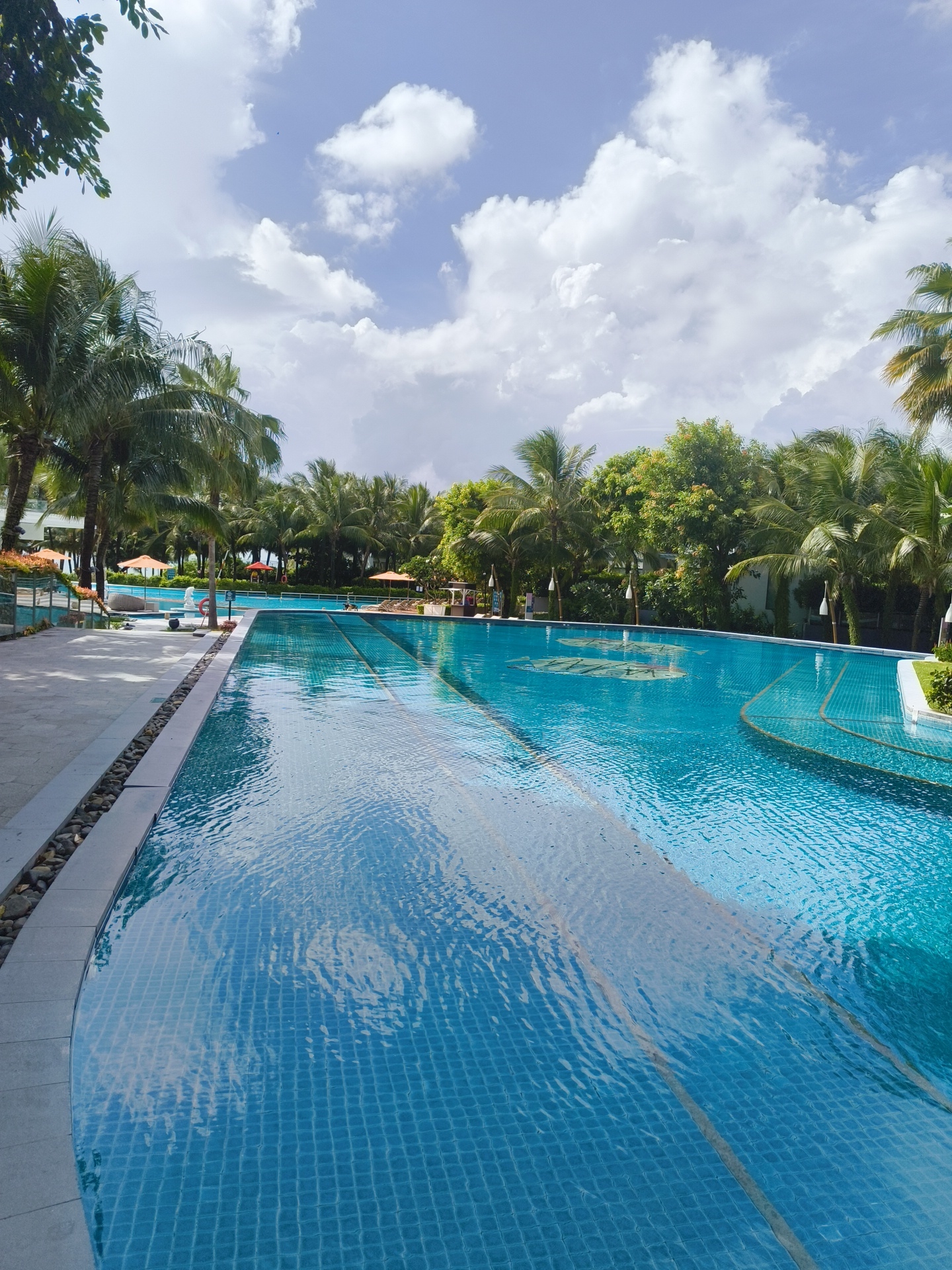 The image depicts a serene swimming pool area at a resort in Phú Quốc, Vietnam. The foreground is dominated by a large, clear blue swimming pool with tiled bottom, reflecting the sky and surrounding palm trees. The pool has distinct lanes marked with darker tiles, suggesting it is designed for lap swimming.
Along the left side of the pool, a paved walkway lined with small, dark stones separates the pool from a shaded area with lounge chairs and orange umbrellas. Beyond this, a building with white walls and visible orange umbrellas can be seen, hinting at guest accommodations or facilities.
The background is lush with a dense arrangement of tall palm trees and other tropical foliage, creating a verdant backdrop against a bright, partly cloudy sky. The clouds are puffy and white, characteristic of a tropical climate. The overall impression is one of a peaceful, luxurious resort setting designed for relaxation and enjoyment under a sunny sky.