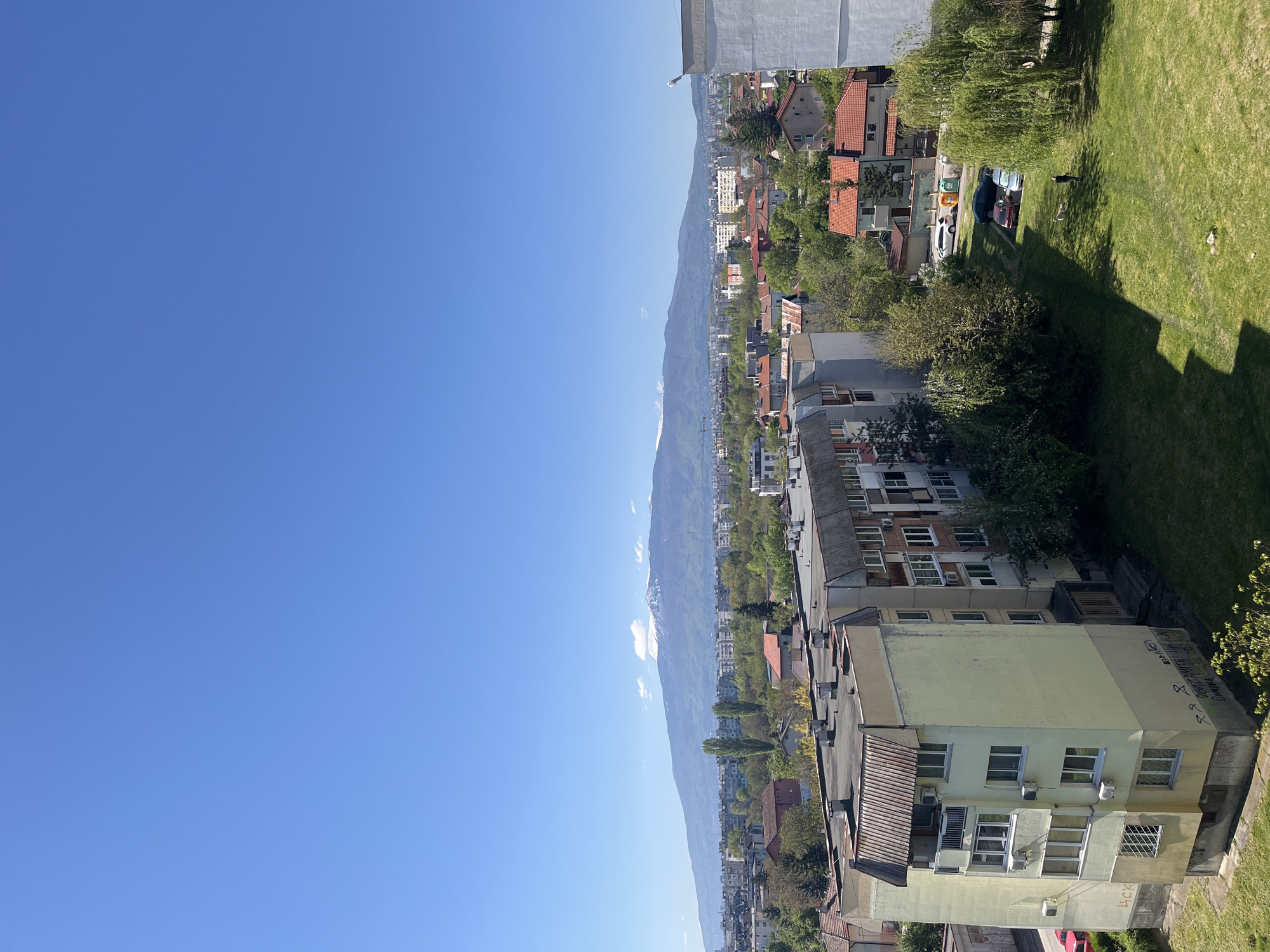 A high-angle shot depicts a city view in Sofia, Bulgaria, under a clear blue sky. Buildings of various heights and architectural styles dominate the foreground, ranging from older, low-rise structures to taller apartment blocks. The buildings are interspersed with trees and greenery. A long mountain range extends horizontally across the midground. In the right foreground, a grassy area is visible with a few trees and some parked cars.