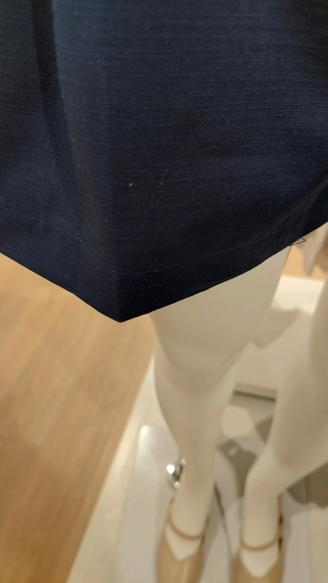 The image is a close-up shot of a dark blue fabric garment, possibly a dress or skirt, displayed on a mannequin. The fabric has a visible texture with fine horizontal lines. A hemmed edge is visible in the lower left quadrant of the image, with a neat stitch line. Below the garment, parts of a white mannequin's legs and beige ballet flats are visible. The mannequin is standing on a white surface, possibly a display stand. In the background, a light brown wooden floor is partially visible. The overall lighting is bright, suggesting an indoor retail or display environment. The image does not contain any recognizable landmarks or context to place it within the city of Bekasi, Indonesia.