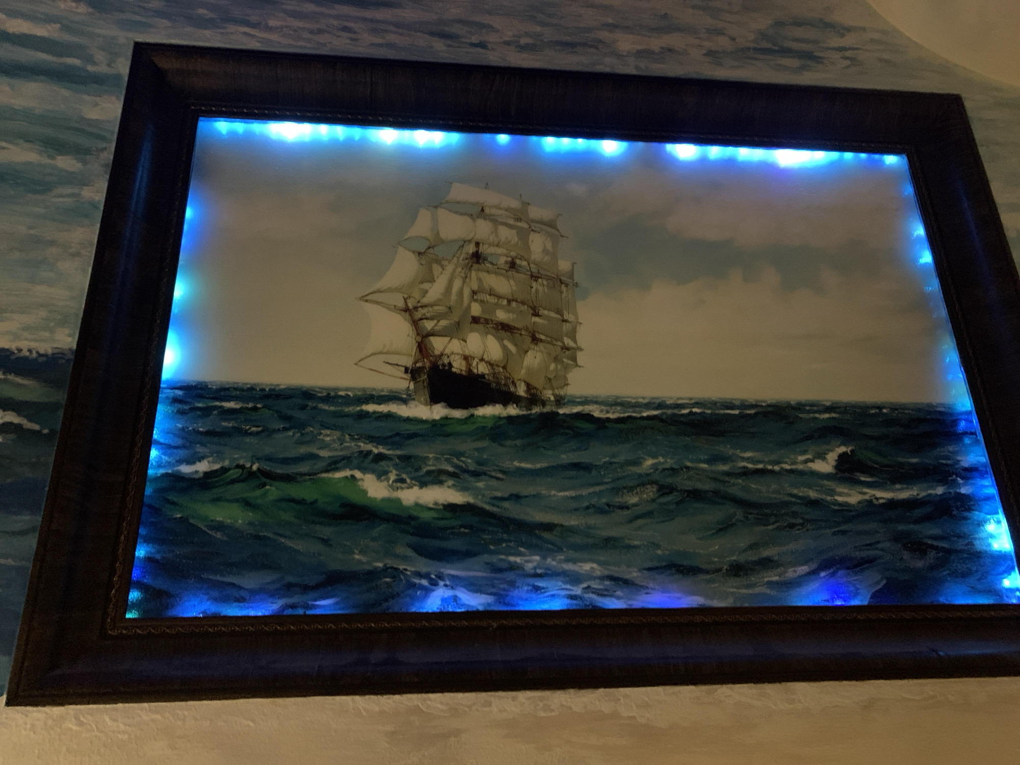 The image shows a framed painting of a tall ship at sea, illuminated with blue lights around the inner edge of the frame. The frame is dark brown and ornate. The painting depicts a large sailing ship with multiple sails cutting through choppy, blue-green water. The sky in the painting is cloudy and muted. The painting is mounted on a wall with a seascape mural partially visible in the background. The location is unknown; there is no data or any elements in the image to show that this image was taken in the city of Severodvinsk, Russia.