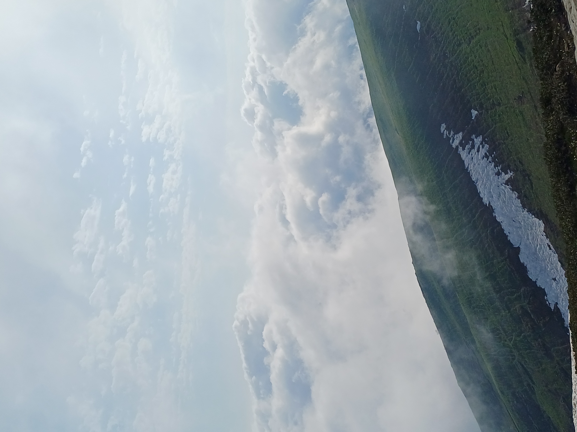 The image shows a panoramic view from a mountainside, likely in Estosadok, Russia. The foreground features a steep, green mountain slope dotted with patches of lingering snow. wisps of mist drift across the grassy terrain. In the midground, a vast expanse of white, fluffy clouds stretches out, creating a sea of clouds that obscures the lower landscape. Above the clouds, a pale blue sky is visible, with scattered, wispy white clouds. The lighting suggests it is daytime, possibly late morning or early afternoon. There are no visible people or man-made structures in the immediate vicinity, emphasizing the natural beauty of the mountain environment.