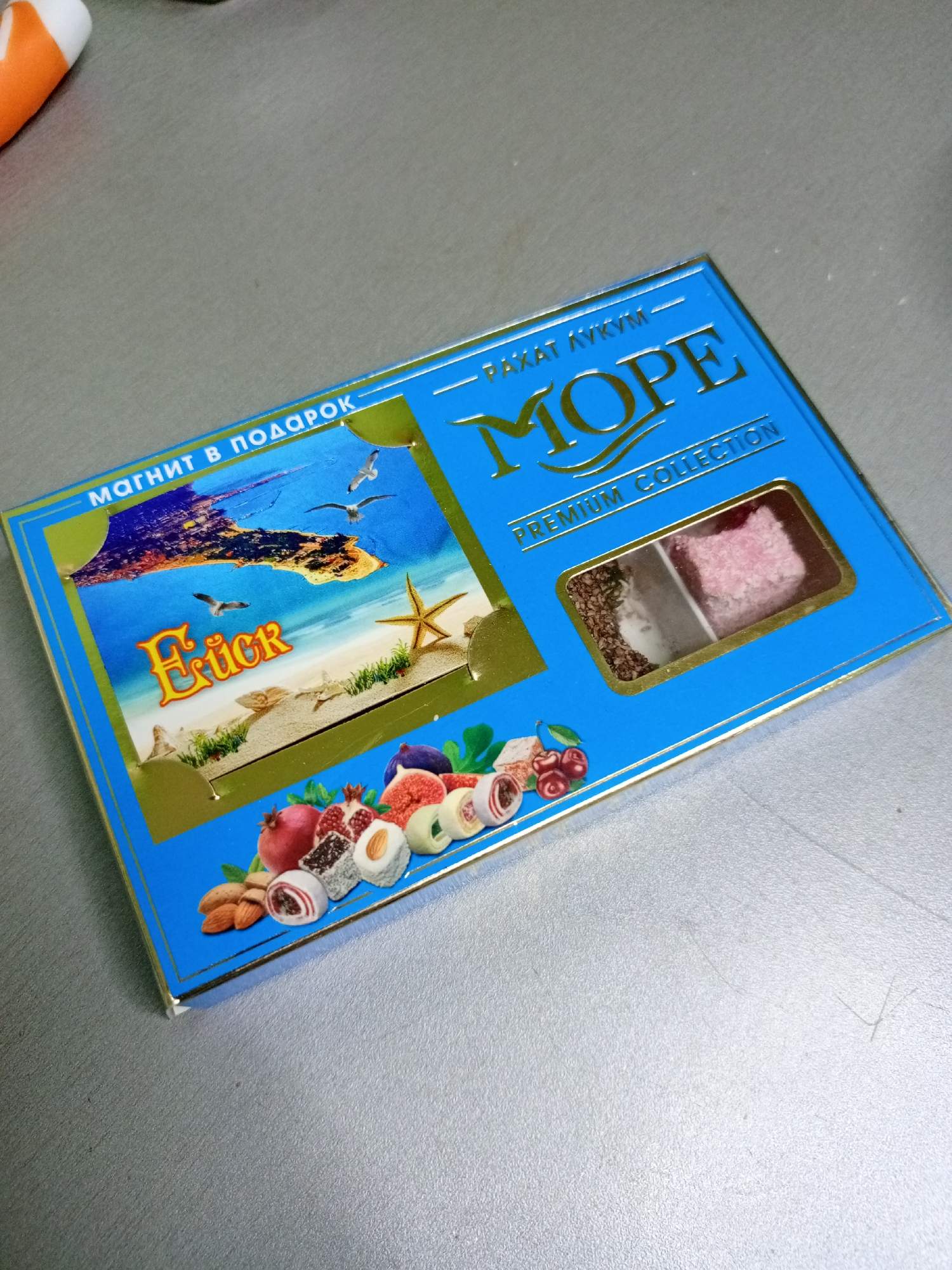 An overhead shot shows a blue box of sweets with a beach scene on it, positioned on a grey metallic surface. The box is angled with the top left corner closer to the viewer. The box features gold text and illustrations. On the left side, in gold lettering, it says "МАГНИТ В ПОДАРОК" (Magnet as a gift), followed by a beach image with seagulls and the word "Ейск" (Yeysk) in a stylized font. The top of the box has gold lettering that says "— РАХАТ КУКYМ —" (Rachat Küküm), and "MOPE" (Sea) in large gold letters. Below "MOPE" it says "PREMIUM COLLECTION" in smaller gold letters. There is a rectangular window in the box showing two pieces of confectionary; one is a dark square covered in nuts or seeds, and the other is a pink square covered in a white granular substance. The bottom of the box depicts an assortment of fruits and nuts, including almonds, pomegranates, cherries, and what appear to be figs, alongside several other sweets. The overall impression is of a product shot for a food item.