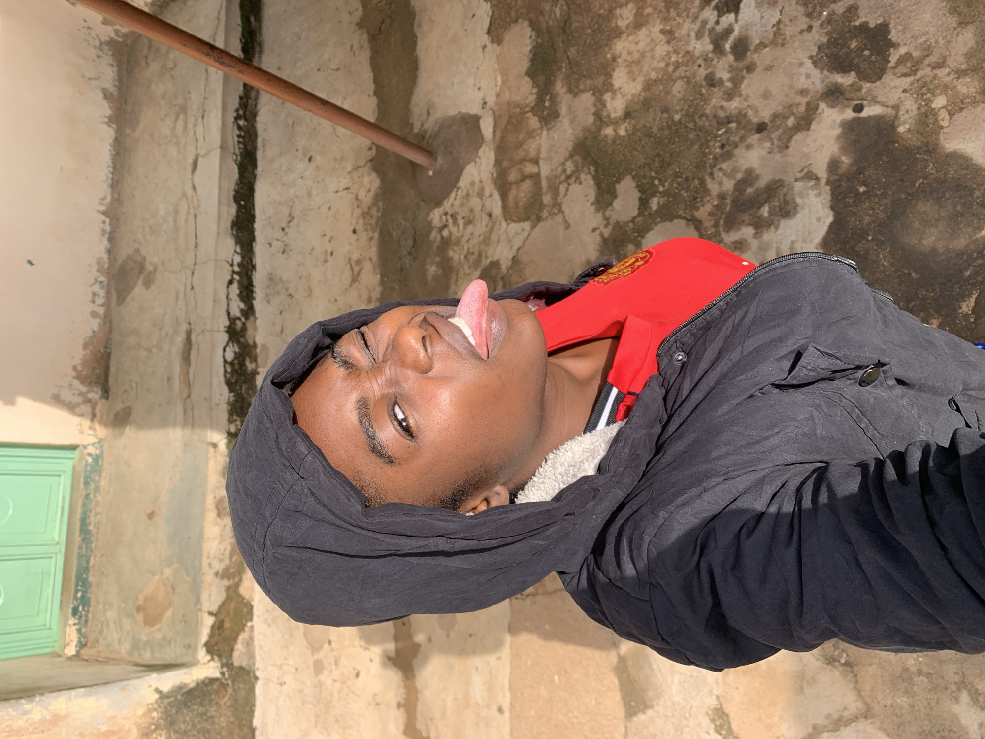 The media file shows a young Black person with their tongue sticking out, wearing a red shirt and a black hooded jacket. They are positioned in what appears to be an outdoor setting with a weathered, concrete-like ground and a textured wall in the background. A copper pipe runs diagonally across the upper left portion of the image. To the left, a light green door with two panels is visible, set against a lighter-colored wall. The lighting suggests it is daytime, with shadows indicating the sun is present. The person's expression and gesture, with their tongue out, could be interpreted as playful or humorous. The overall scene seems to be in a somewhat rustic or unrefined environment, possibly a street or an alleyway. The visible textures of the ground and walls suggest an outdoor location in Rafin Par, Nigeria. No visible text is present in the image.