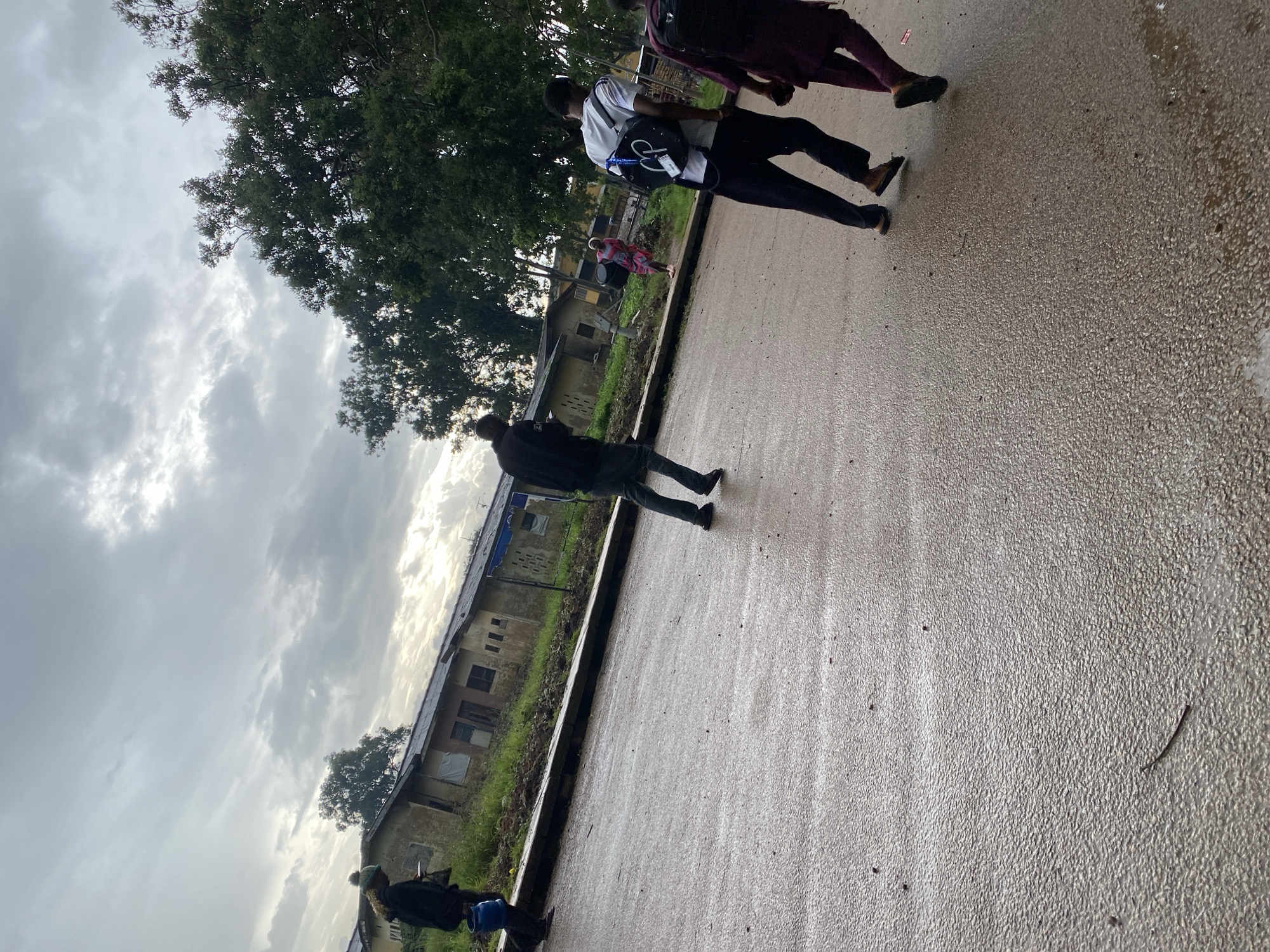 In Rafin Par, Nigeria, on a wet asphalt road, a group of four individuals are walking. The sky is overcast with a mix of light and dark clouds. To the left of the road, there is a grassy area with several small, weathered buildings that appear to be residential structures, some with visible windows and doors. In the background, dense green trees line the scene.
The individual furthest to the left is carrying a blue bucket and wearing a dark jacket and a light-colored beanie. Next to them, a person in a black jacket and dark pants is walking away from the camera. Further down the road, a person in a white t-shirt and dark pants, carrying a black backpack, is walking towards the right. The individual closest to the camera on the right is wearing a dark maroon outfit and carrying a black backpack, also walking towards the right. A woman in a patterned dress carrying a black bucket is walking in the middle ground, between the buildings and the road. The road surface is wet, with visible reflections of the sky and surrounding elements.