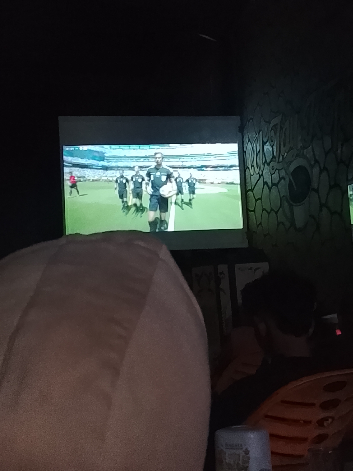An image captures a dimly lit room where a projection screen displays a football match. Several figures in black and white uniforms are visible on the screen, with one individual holding a football. In the foreground, a person is partially visible, their head and shoulder covered by a light brown fabric, suggesting they are seated or leaning. To the right, another individual's head and shoulders are seen, looking towards the screen. An orange plastic chair with a distinctive pattern is also visible. The background wall features a textured pattern resembling scales or stones, with some visual elements that could be art or decor. The overall ambiance suggests a casual viewing environment, possibly a bar or a common area. The specific location in Rhieng Blang, Indonesia, is not visually discernible from the image itself.