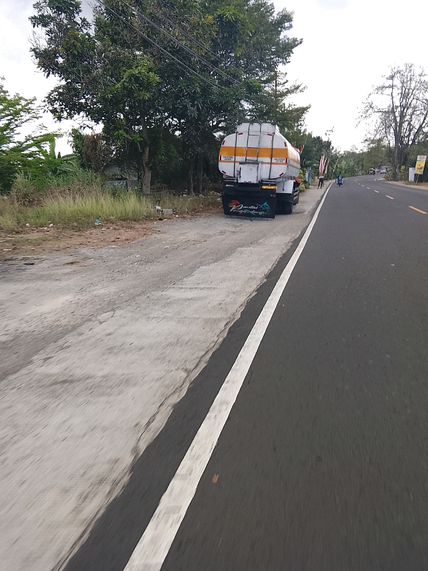 The image depicts a road scene in Kertahayu, Indonesia, on a cloudy day. A large white tanker truck with yellow stripes is parked on the side of the road, which is asphalt. The truck has "Pantai Pangandaran" written in stylized lettering on its rear mud flaps. To the left of the truck, there is a grassy area with tall dry grass and some debris. Beyond the grass, a dense cluster of trees and vegetation is visible. Further down the road, two people are seen, one standing and the other on a blue motorcycle. There are other vehicles and trees in the distance. The road has a white dashed line marking its edge and a solid yellow line down the center. The overall environment suggests a rural or semi-rural setting.