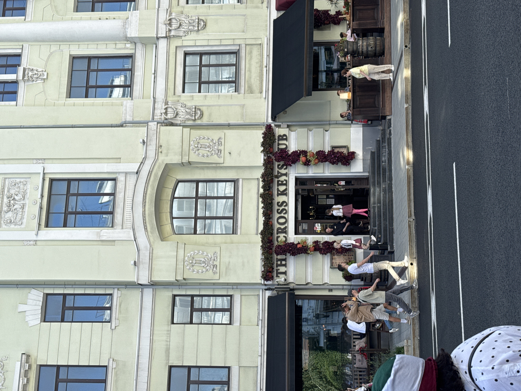 This is a photograph taken from a street-level perspective, looking towards the entrance of "THE CROSS KEYS PUB" in Moscow, Russia. The building facade is a pale green with white decorative elements. The pub entrance is centered, with stairs leading up to it. Above the entrance, a sign reads "THE CROSS KEYS PUB". Red and green garlands are draped around the entrance and along the building's facade, suggesting a festive or seasonal decoration.
Several people are visible in the scene. On the sidewalk in front of the pub, a group of individuals are standing and walking. A woman in a light-colored outfit is walking towards the pub, while a man in a white t-shirt and beige pants is walking away from it. Two people, a man in a light brown jacket and shorts and another person in a beige t-shirt and gray pants, are walking on the sidewalk. Near the pub entrance, a person in a white top and pink skirt is standing, and two other individuals in dark clothing appear to be employees.
To the left of the pub entrance, an outdoor seating area with tables and benches is visible, partially obscured by a black awning. Several patrons are seated at tables in this area. To the right of the pub, a bar area with patrons is visible, with wooden barrels and seating. A woman in a yellow top and beige pants is standing near this bar area.
The street in the foreground has a dark asphalt surface with white lane markings. A person wearing a distinctive hat with red and white trim is partially visible in the bottom left corner of the image, looking towards the street. The sky is overcast.
