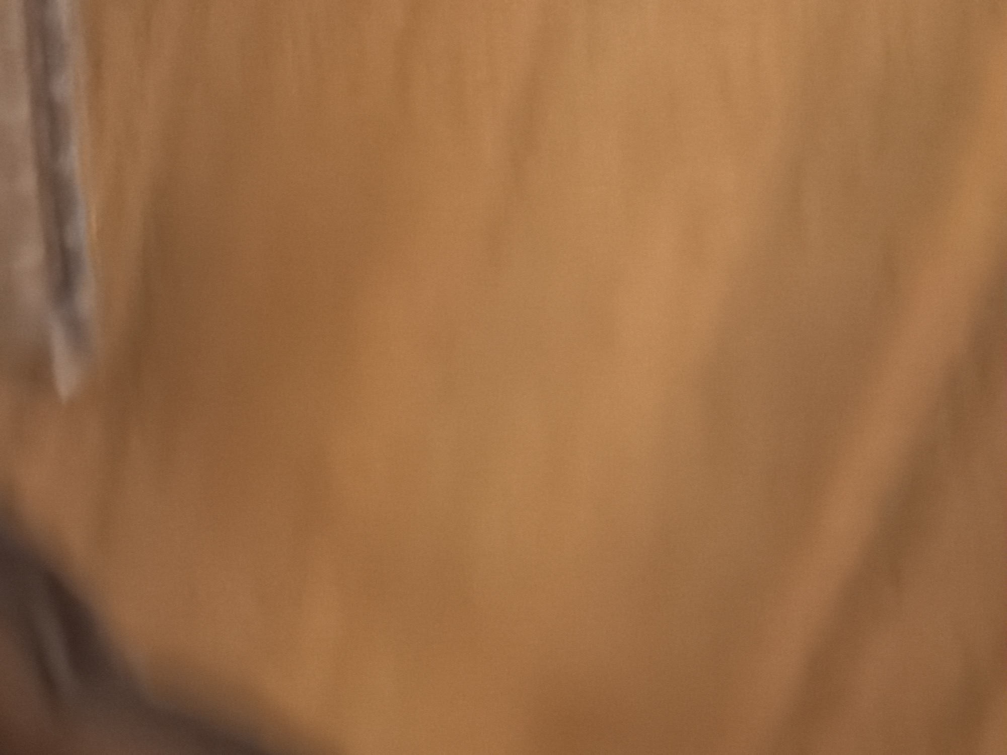 The image shows a close-up, blurred view of a brown, textured surface. The lighting creates subtle highlights and shadows, suggesting a fabric or material with a slight sheen and possibly some wrinkles or folds. There are no discernible objects, people, or environmental details visible in the frame.