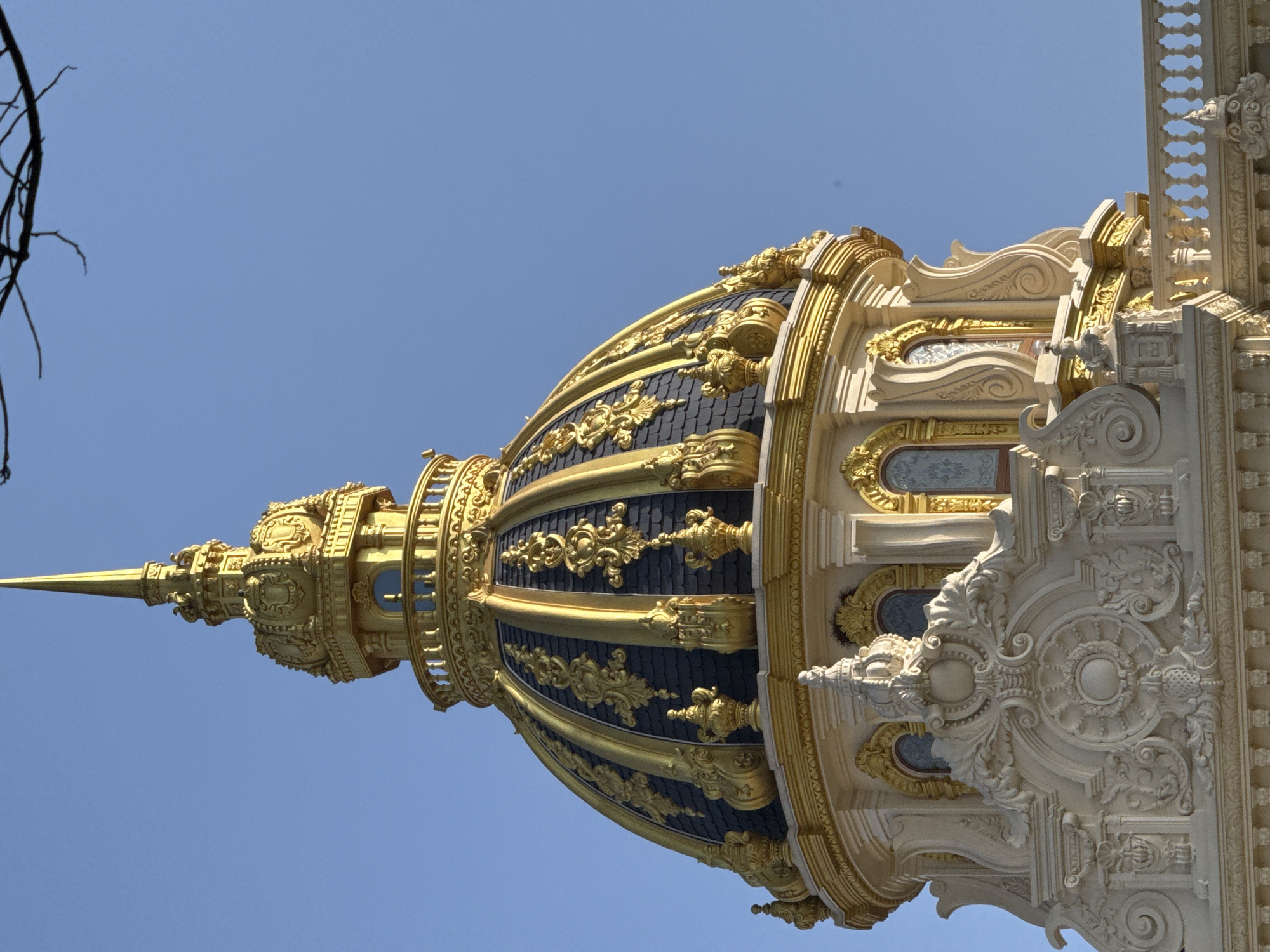 The image displays a close-up view of a golden dome and ornate architectural elements of a building against a clear blue sky. The dome is segmented and adorned with intricate golden scrollwork over dark blue tiles. Below the dome, a white stone facade features detailed carvings, including a circular motif with a central sphere, scroll patterns, and decorative elements resembling crowns and foliage. Small arched windows with decorative gold trim are visible on the building's structure. A dark tree branch extends from the top right corner of the frame. The building appears to be a significant landmark, possibly a palace or religious structure, characterized by its opulent and detailed design. This building is the Dinh Doc Lap or Independence Palace, located in Ho Chi Minh City, Vietnam, not Da Lat.