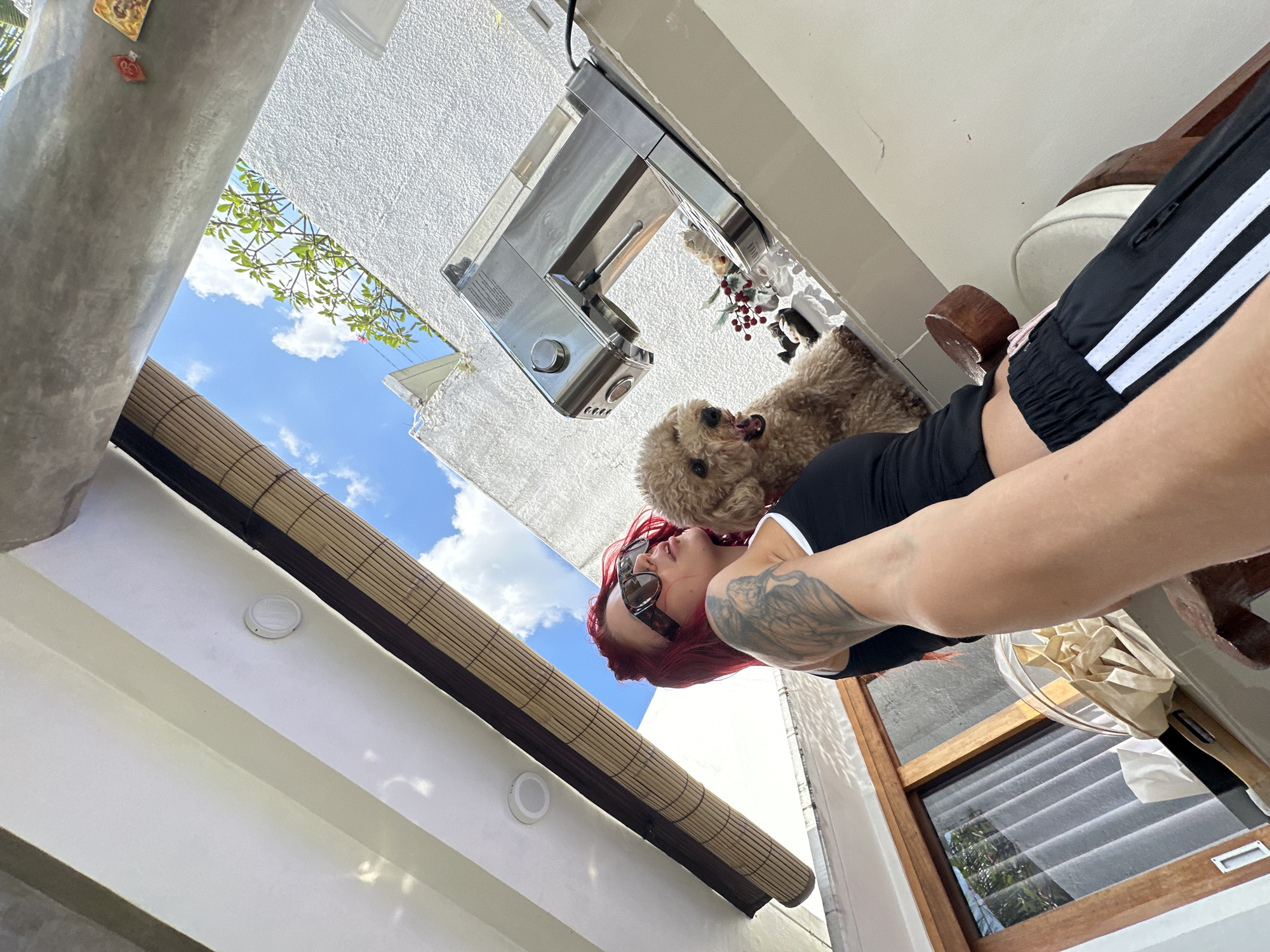 In North Kuta, Indonesia, a woman with bright red hair and sunglasses poses with a small, light brown poodle. She is wearing a black tank top with white trim and black athletic pants with white stripes. The dog is perched on a white ledge next to a stainless steel coffee maker. Behind them, a bright blue sky with fluffy white clouds is visible, partially obscured by bamboo blinds and the building's white textured wall. The overall scene suggests an outdoor or semi-outdoor setting, likely a balcony or patio.