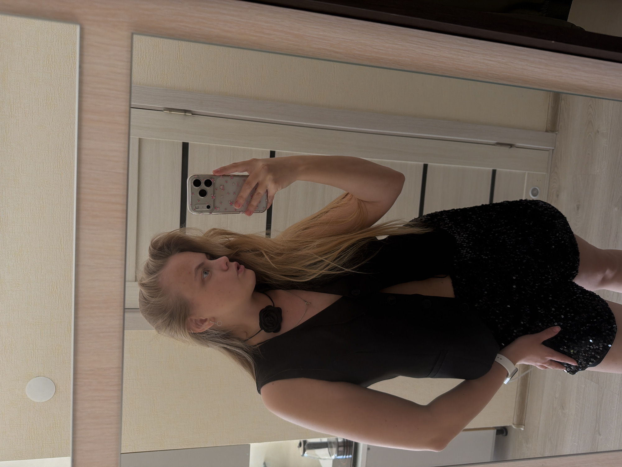 A woman with long blonde hair is taking a selfie in a mirror. She is wearing a black sleeveless dress and a black flower choker. Her dress appears to be decorated with sequins. She is holding a smartphone with a floral case. The background shows a light-colored door with vertical paneling and a wall. The mirror reflects part of a bathroom sink area. This image doesn't contain sufficient information to confirm the location is Izhevsk, Russia.