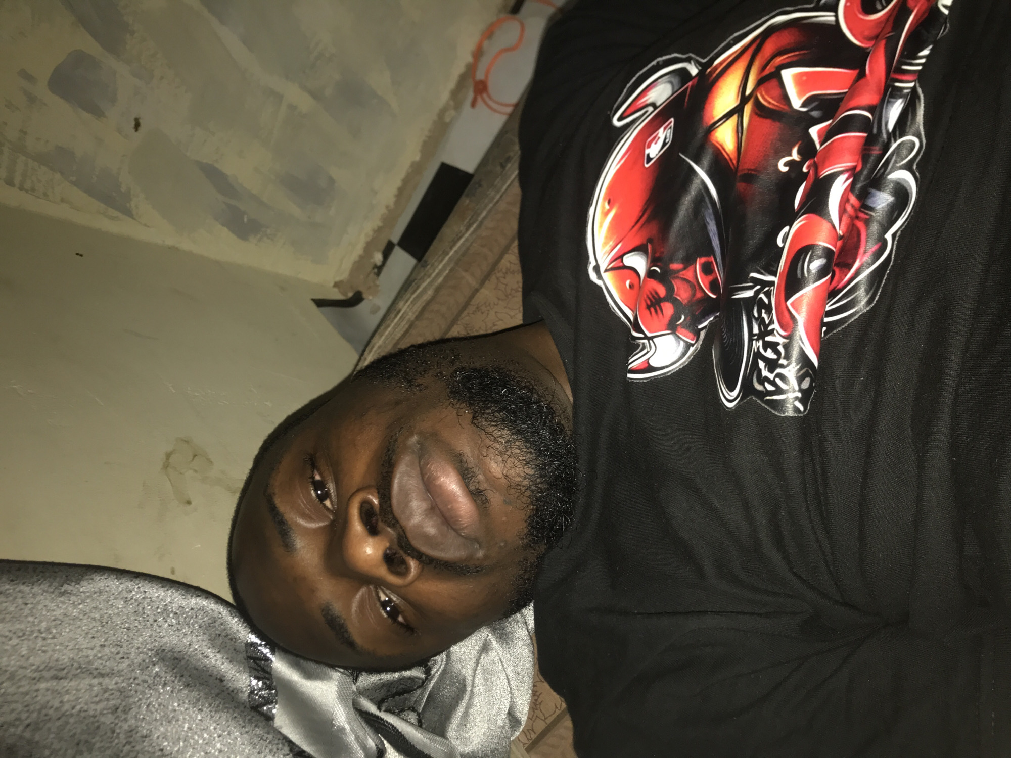 An adult dark-skinned male is depicted in a close-up, upward-facing shot, lying on his back within an indoor setting. He possesses a short beard, mustache, and dark hair, looking directly into the camera with open eyes and full lips. He wears a black short-sleeved t-shirt featuring a large, stylized graphic of a red and black bull with white horns and outlining on the chest, which includes a small NBA logo. To the left of his head, a crumpled mass of shimmering grey fabric is visible. The background consists of a light-colored wall with visible brushstrokes and irregular, darker marks or stains. Below his right arm, a section of brown patterned fabric is present, and further to the right, a black and white checkered pattern can be observed, along with a loosely coiled orange cord. The lighting suggests an overhead source. The location is in the city of Unknown, Unknown.