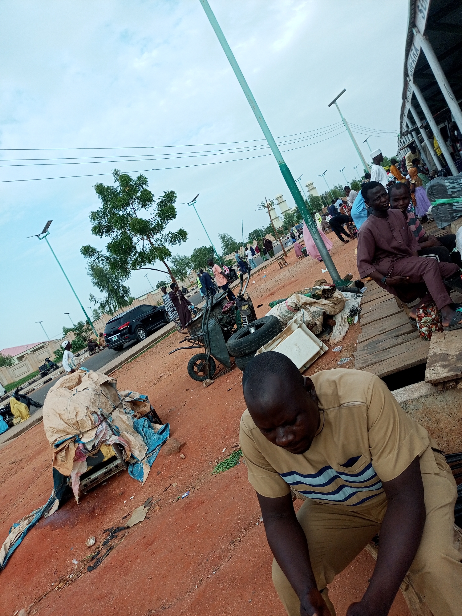The image depicts a street scene in Gombe, Nigeria, likely a market area, characterized by red earth ground and an overcast sky. In the foreground, a man in a tan outfit with blue and white wavy stripes is seated, looking down. To his left, a large pile of covered items, possibly goods or refuse, is present. A dark-colored SUV is visible on the road behind the pile.
Further back, a variety of people are seen in the background, some walking, others sitting. One individual, dressed in a brown outfit and a white cap, is standing near a cart. A partially visible market structure with white pillars is to the right of the image. Several streetlights with solar panels are also scattered throughout the scene. The overall impression is one of a busy, everyday urban environment.