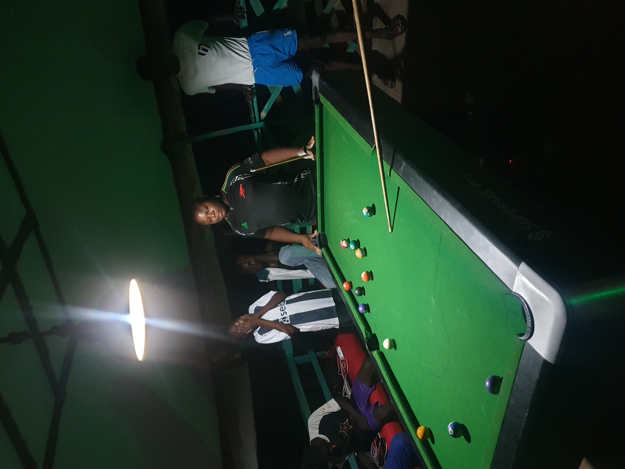 The image depicts a group of young people engaged in a game of pool at night. The scene appears to be an outdoor covered area, possibly a makeshift establishment, with a green pool table as the central focus. A bright overhead light illuminates the table and some of the players. Several individuals, primarily young men and a few young women, are gathered around the table. One person, a man in a black shirt, is actively playing, holding a pool cue. Others are observing, some with expressions of concentration or amusement. The environment suggests a casual, social gathering. The presence of a "SUPERMAX PRO" logo on the pool table's side indicates the brand of the table. The specific location in Lokoja, Nigeria is not explicitly stated in the image, but the context suggests a local, informal setting.