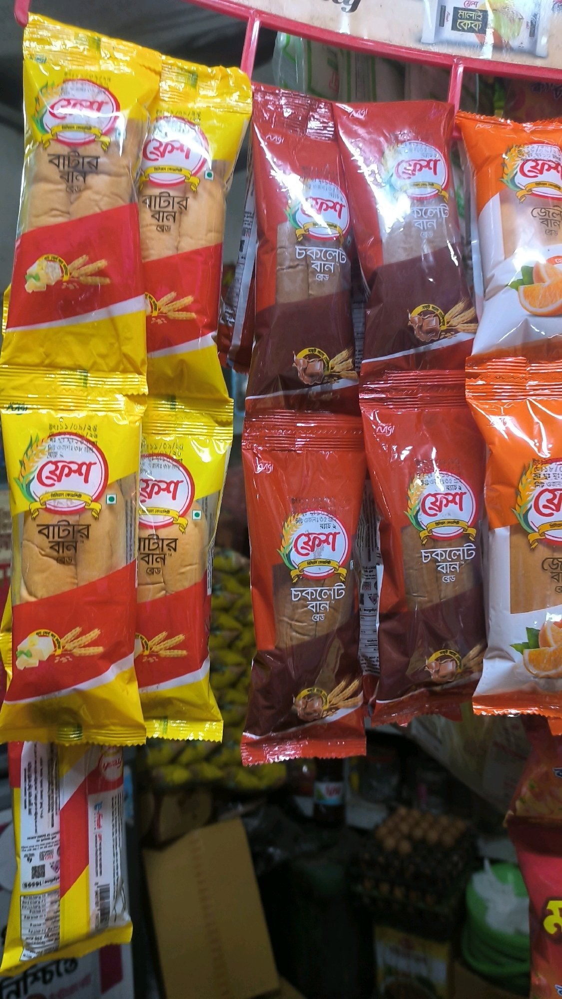 A street vendor's stall in Begumganj Upazila, Bangladesh, displays an array of packaged bread products hanging from a red metal rack. The products are predominantly yellow, red, and orange, with clear plastic windows revealing the bread inside. The branding appears to be in Bengali script. Some packages feature images of wheat stalks and butter or chocolate. In the background, a variety of items are visible, including eggs in a crate, boxed goods, and possibly other food items. The lighting suggests an outdoor or semi-covered market environment.