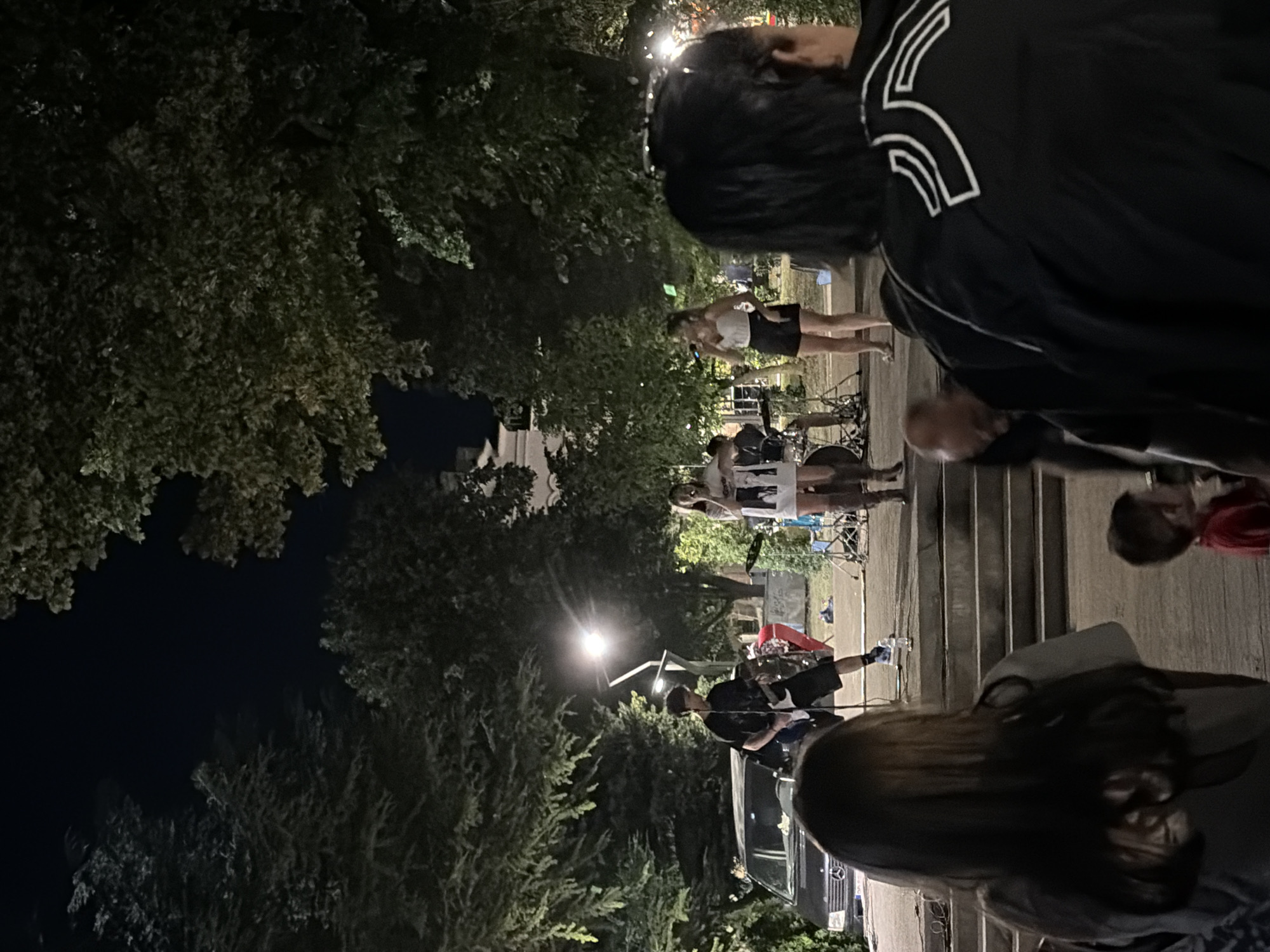 A night-time outdoor concert is taking place, likely in Vratsa, Bulgaria, as indicated by the presence of trees and a building in the background. The stage area features a band with a guitarist and two vocalists, one of whom is actively singing into a microphone. A drum set is visible, and additional band equipment is present. The audience appears to be standing or sitting in front of the stage, with some individuals closer to the front, while others are further back. A dark-colored van is parked near the stage. The scene is illuminated by artificial lighting, including stage lights and streetlights. The overall atmosphere suggests a lively event, possibly a street performance or a small festival.