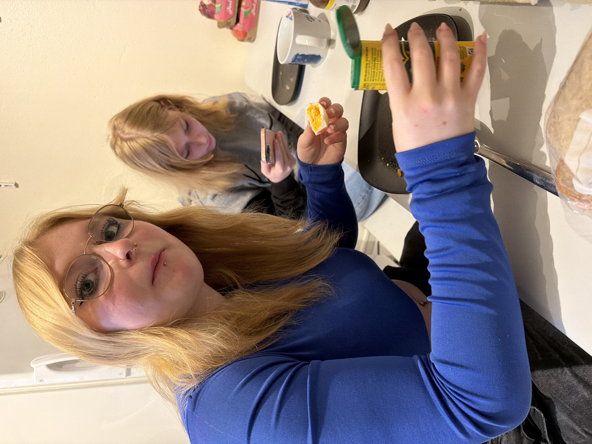 Two young women are seated at a white table, one in a blue long-sleeved shirt and glasses holding a filled egg half and a yellow can. The other, in a gray sweatshirt, holds a cell phone. A black frying pan, a white cup, and a loaf of bread are also on the table. The location of Trassem, Germany, is not discernible from the image.