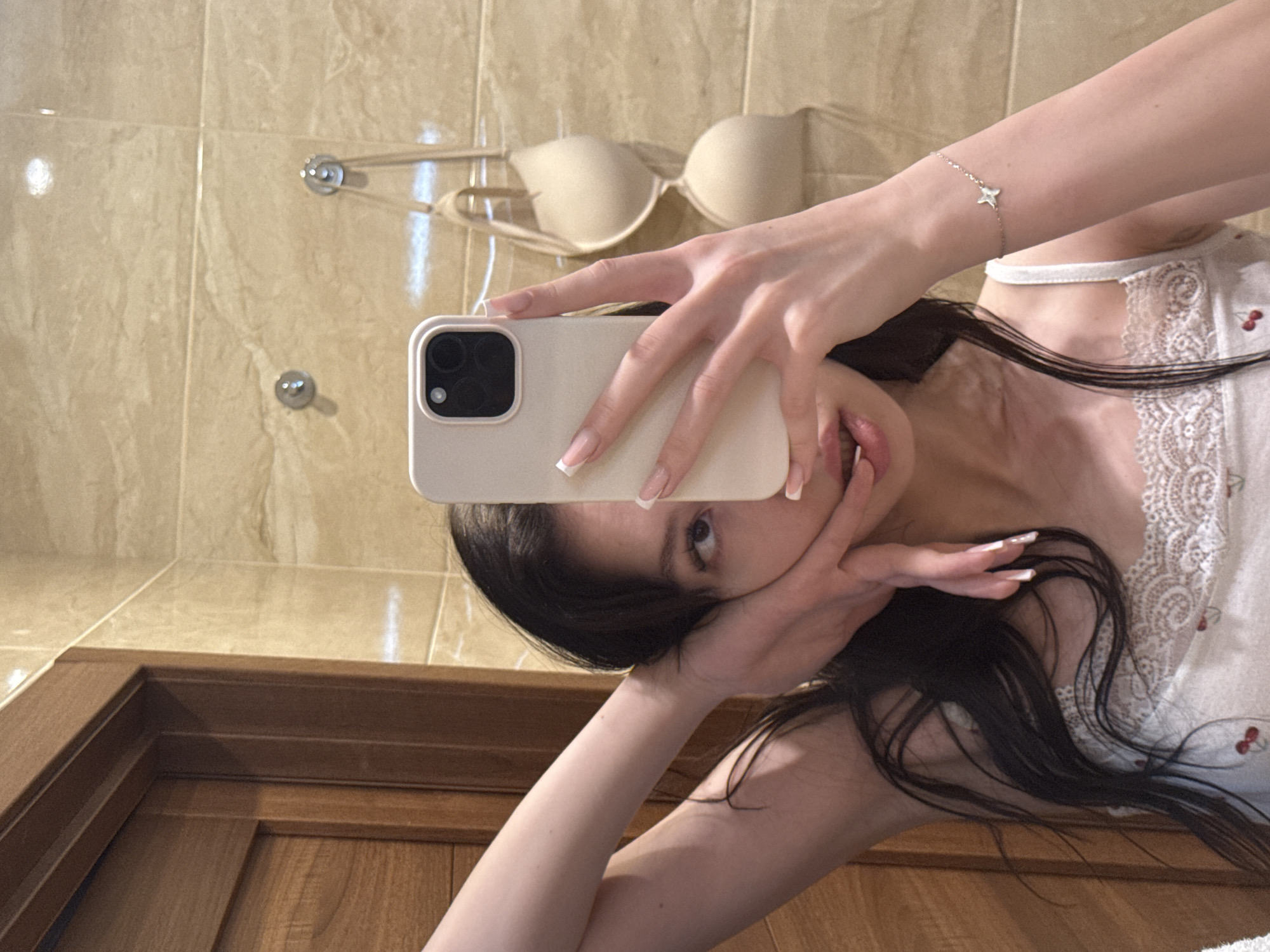 A woman is taking a mirror selfie in a bathroom. She is holding a white phone in her right hand, partially obscuring her face. Her left arm is raised and bent, with her hand resting near her face. She is wearing a white tank top with a lace trim and a cherry pattern. A beige bra is hanging on the wall behind her. The bathroom walls are tiled with a marble pattern. A wooden door is visible to the left of the frame. The image appears to have been taken in a residential bathroom, possibly in the city of Khimki, Russia.